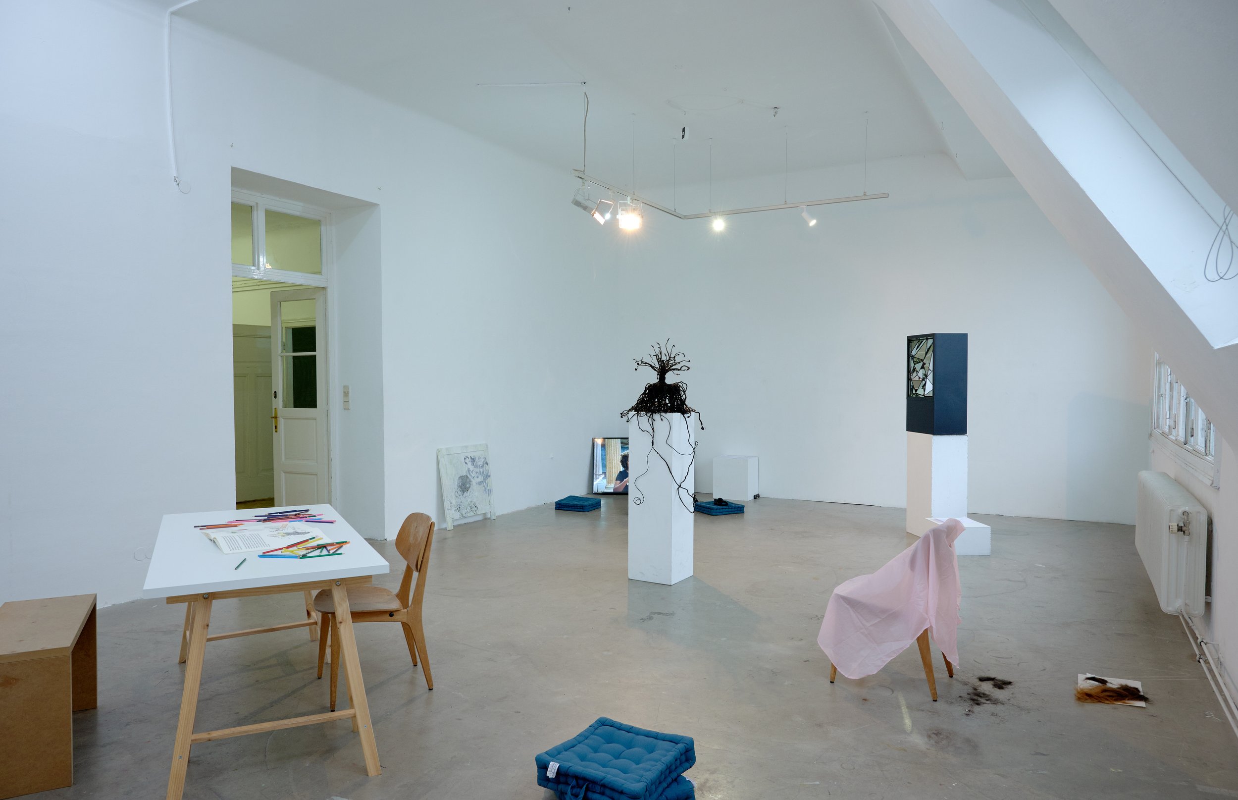 Minimalist art gallery with white walls and wooden floors, featuring sculptures, framed artwork on the floor, and a table with colored pencils.