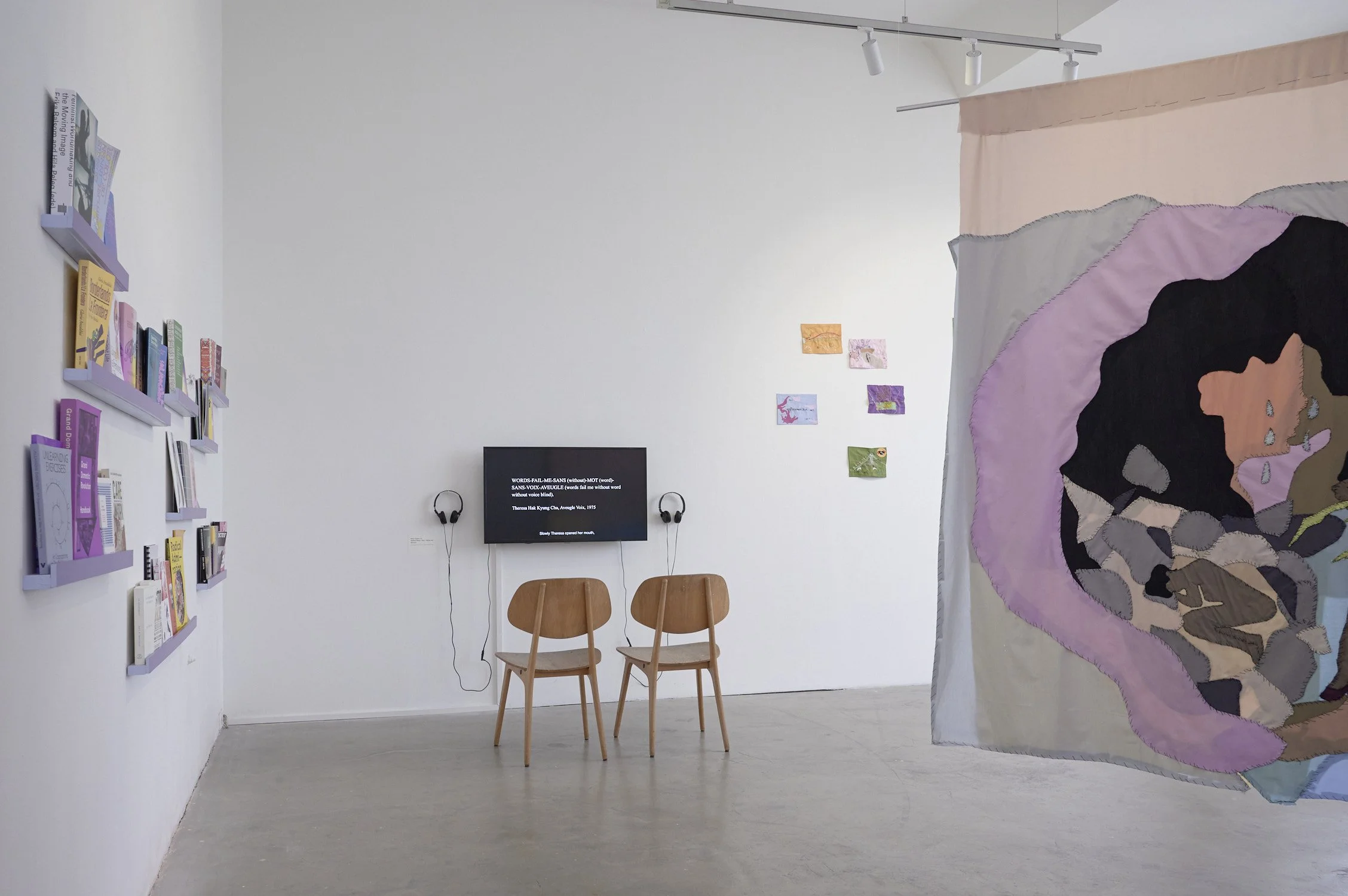 An art gallery with bookshelves, a TV screen, two wooden chairs, and an abstract tapestry.