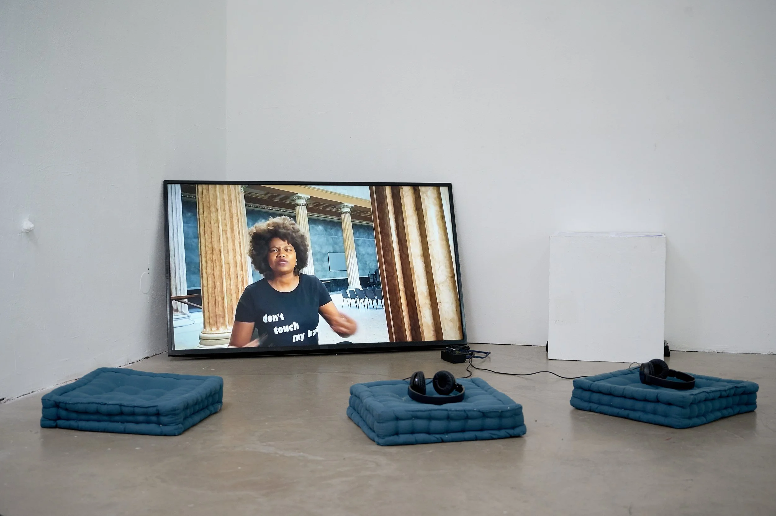 A digital screen displaying a woman with curly hair, standing in an ornate room with gold columns and curtains. In front of the screen are three blue cushions on the floor, each with black headphones placed on top.