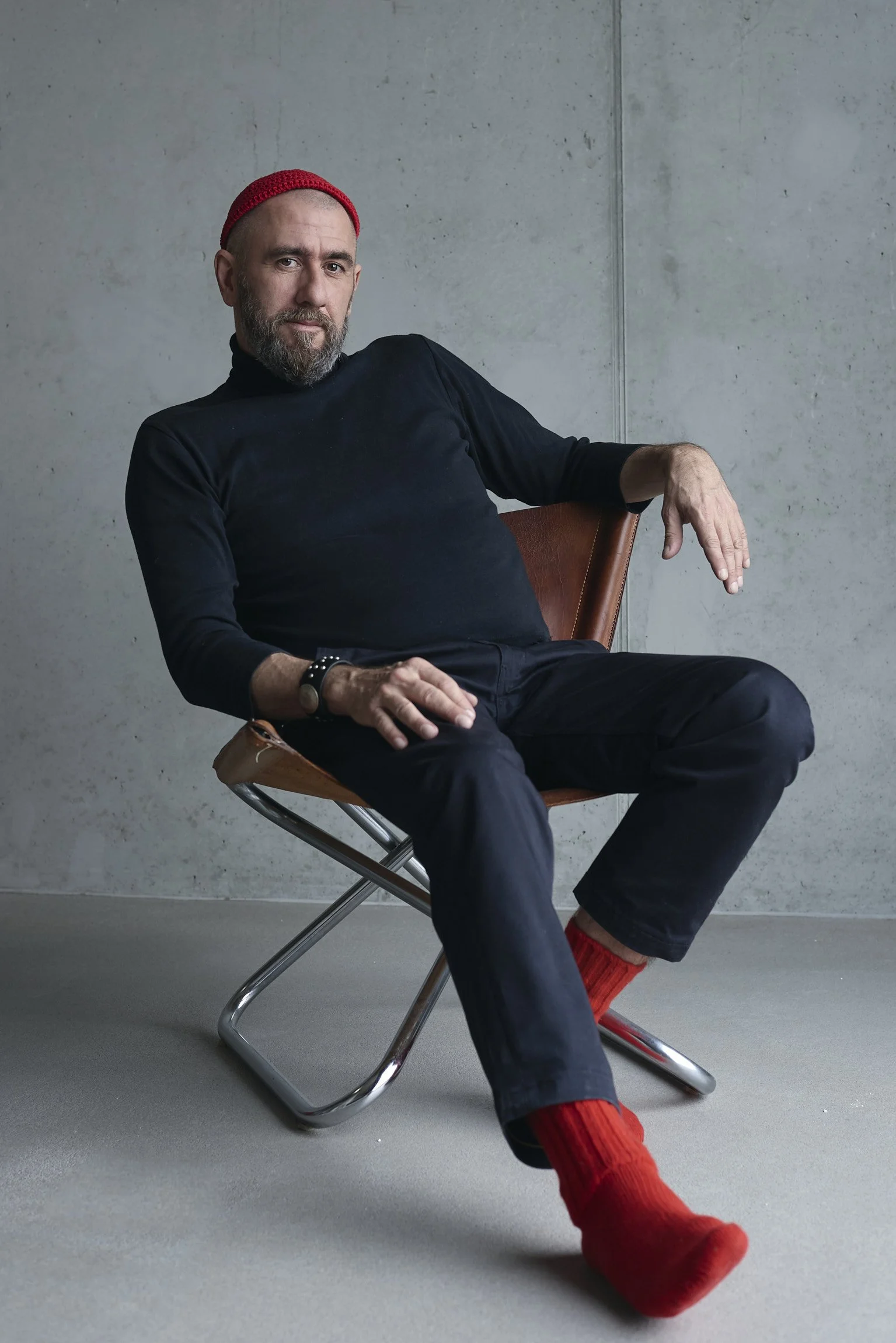 A man with a beard, wearing a red knit cap, black turtleneck, black pants, and red socks, sitting on a brown chair against a concrete wall.