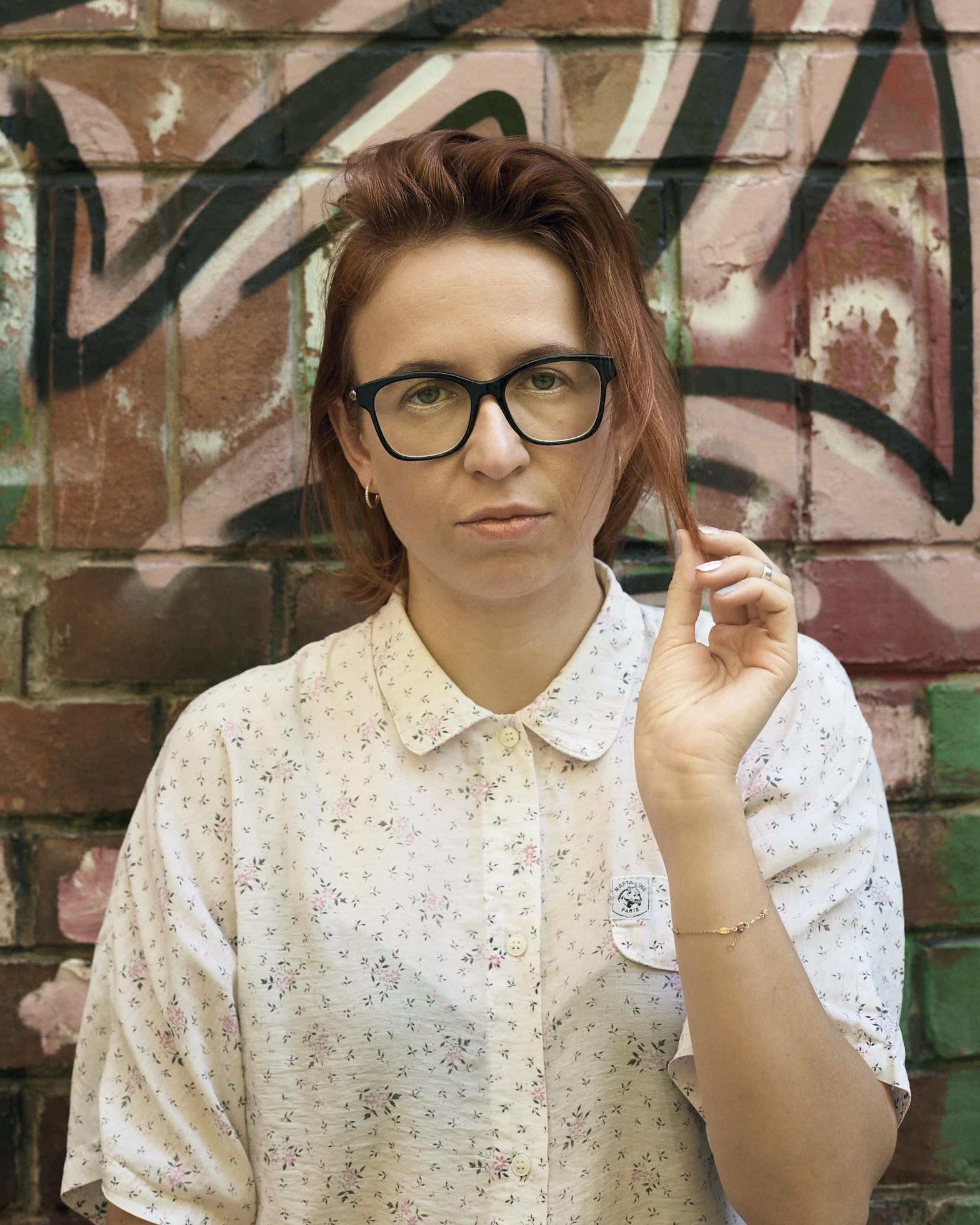 A woman with red hair and glasses standing in front of a graffiti-covered brick wall.