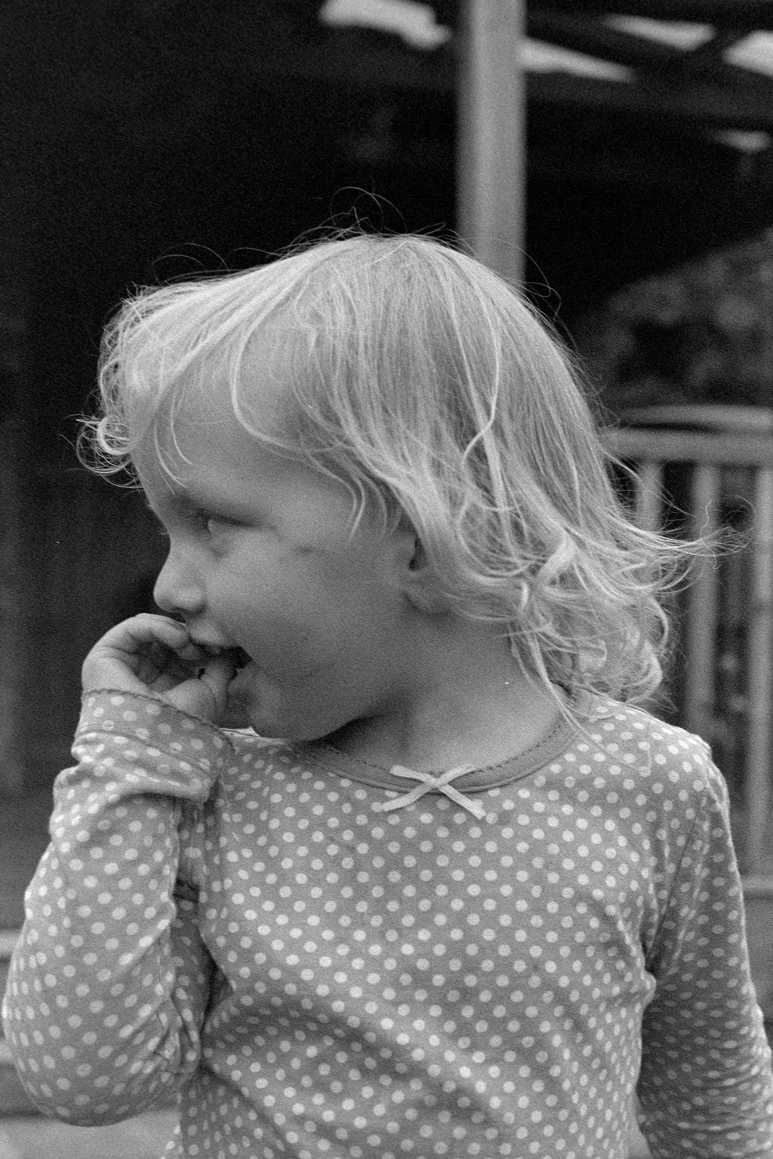 A young girl with light-colored, wavy hair wearing a polka dot long-sleeve shirt with a small bow near the neckline, biting her fingers and looking to the side.