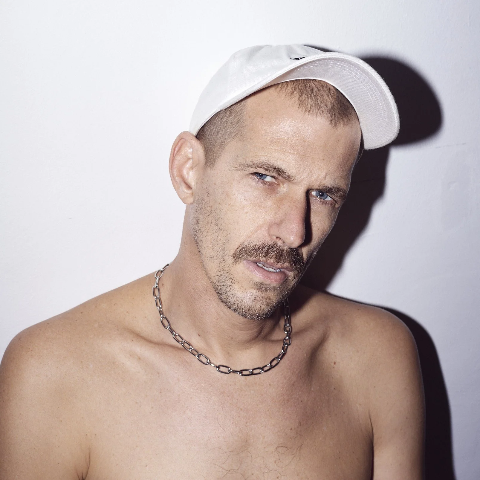 A shirtless man wearing a white baseball cap and a silver chain necklace, looking at the camera with a serious expression, against a plain white background.