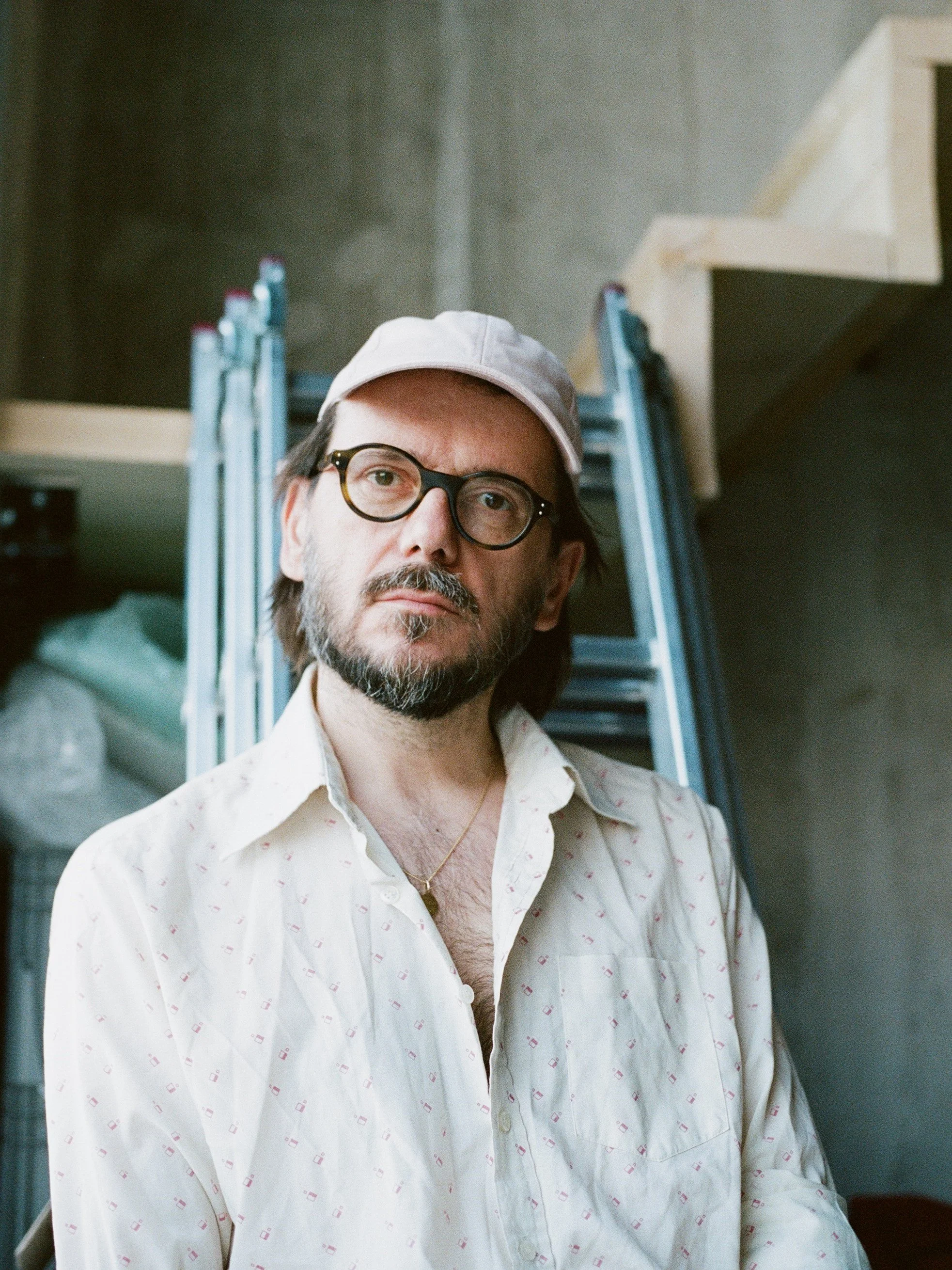 A man with glasses, a beard, and long hair wearing a white cap and shirt with small pattern, standing in front of a ladder and construction materials.