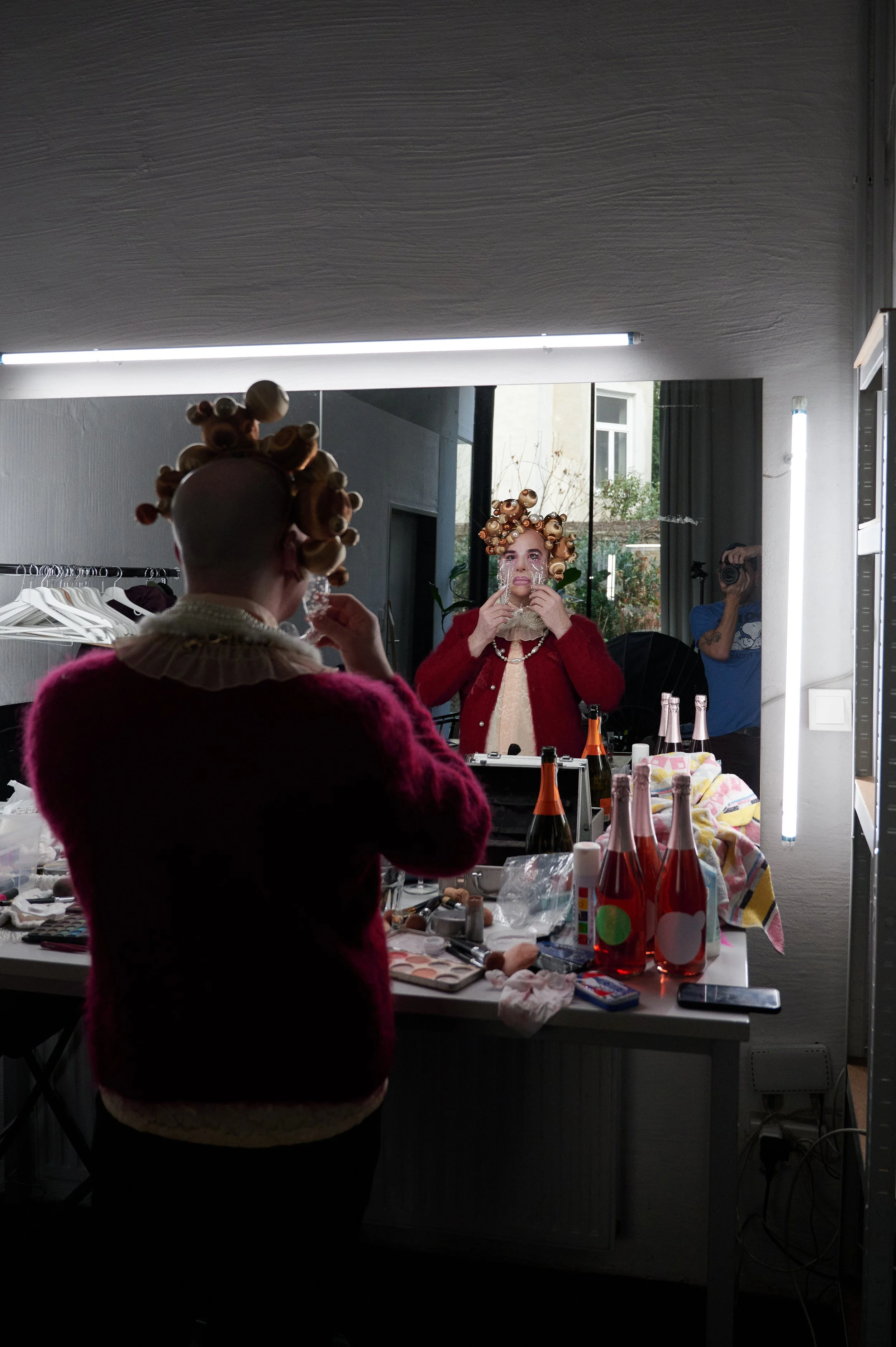 Person applying makeup in the mirror with hair rollers, surrounded by bottles and cosmetics on a cluttered dressing table, reflecting in a large mirror with a photographer in the background.