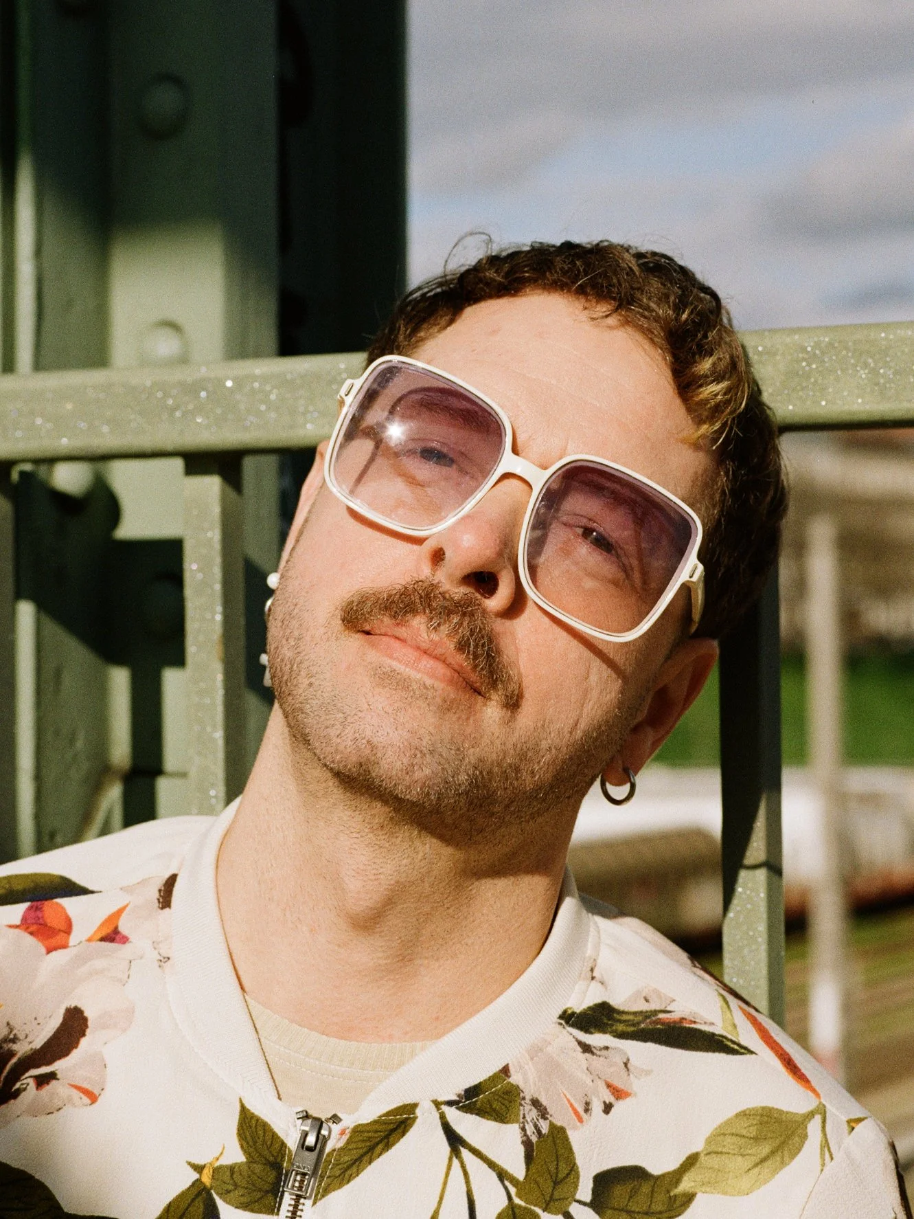 Close-up of a young man with curly hair, a mustache, and earrings, wearing large sunglasses and a floral patterned jacket, leaning against a metal railing outdoors during daytime.