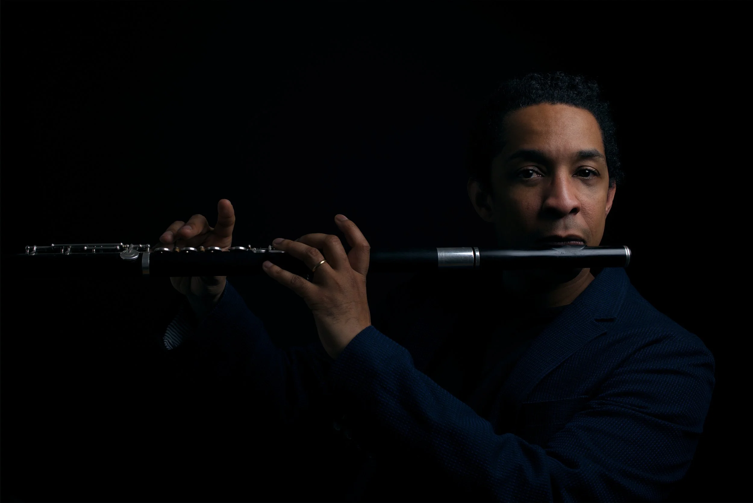 A person with dark hair plays a flute against a dark background, with their eyes looking directly at the camera.