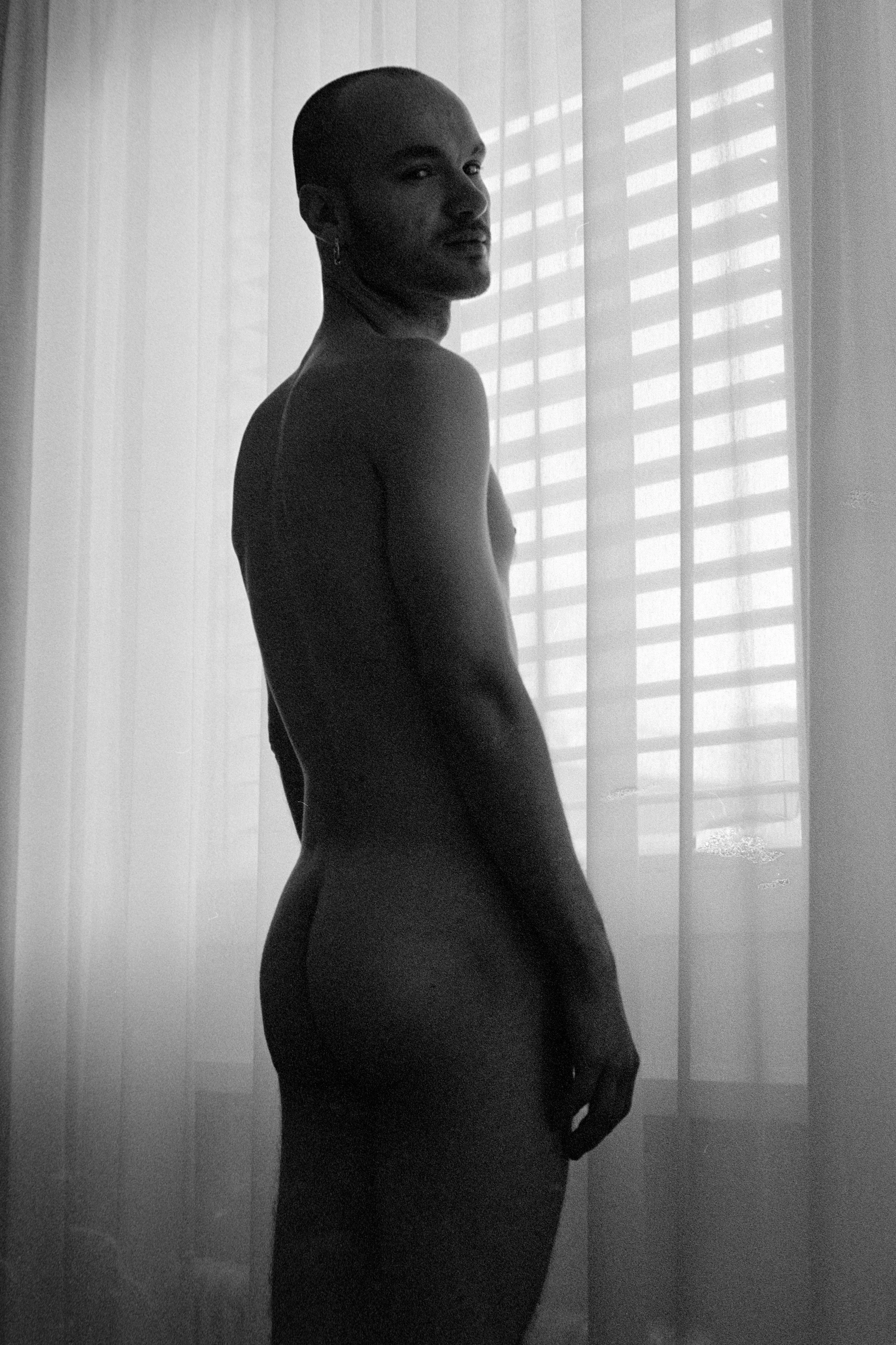 A black-and-white photo of a woman standing near a window with blinds, looking over her shoulder.