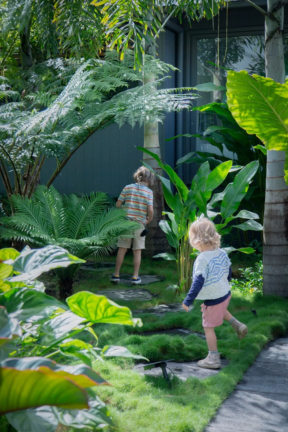 Two children walking on a garden pathway surrounded by lush green tropical plants and trees.