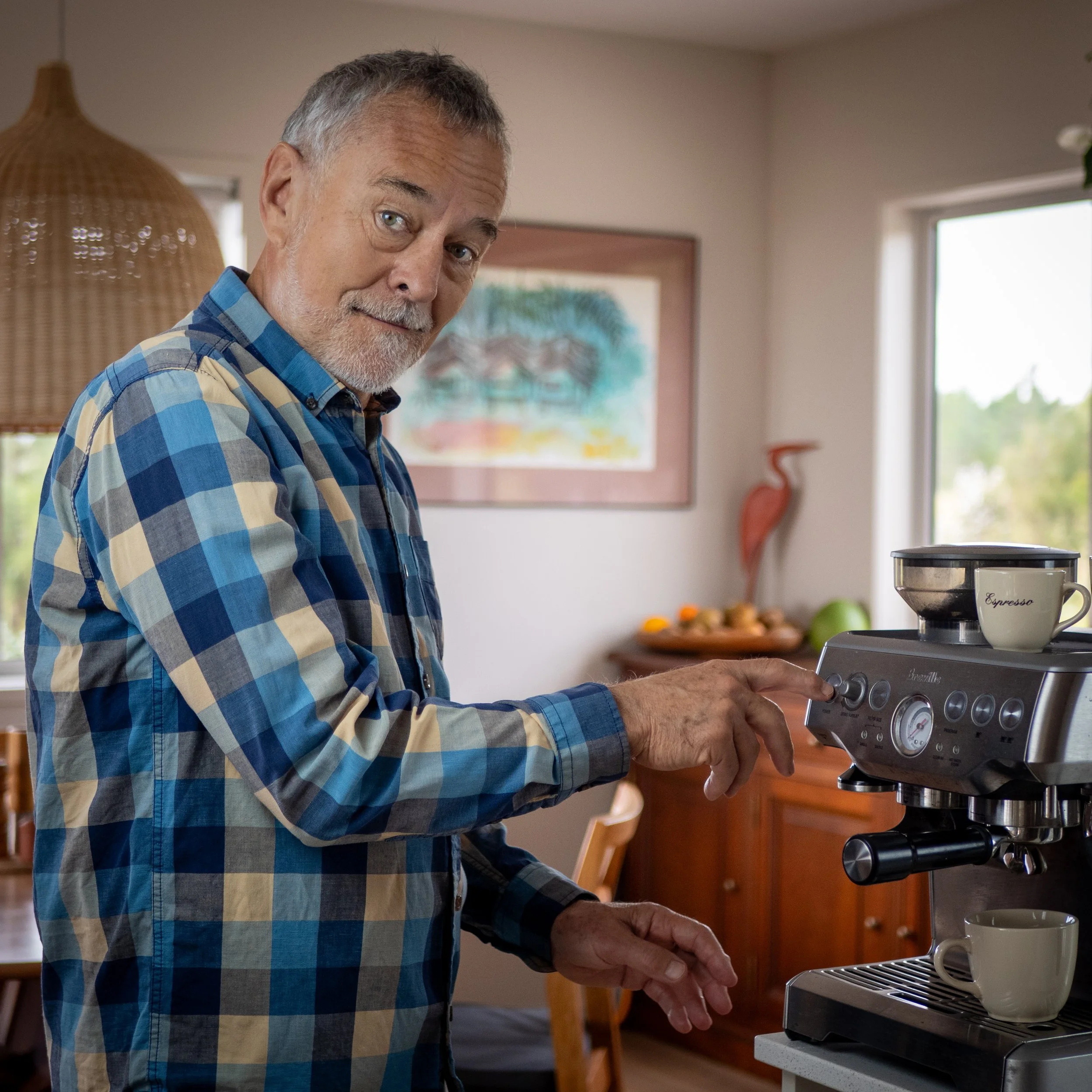 An older man with gray hair and a beard making a coffee in a kitchen. He is pressing a button on an espresso machine, with a coffee mug on top of the machine and another mug placed underneath the spout.