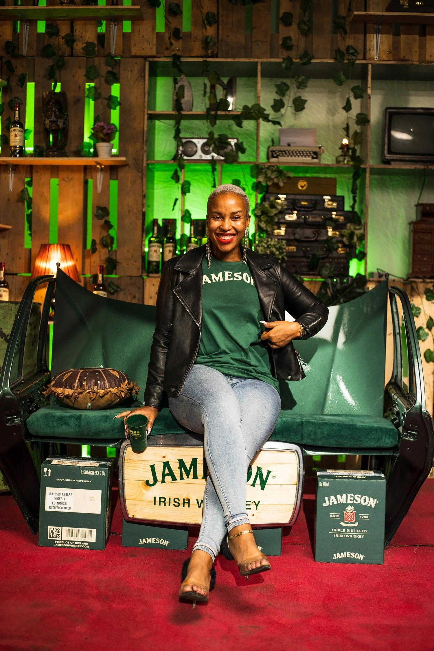 A woman sitting on a vintage green bench decorated with Jameson Irish Whiskey branding. She is smiling and pointing at her shirt, which has 'Jameson' printed on it. The background has a wooden wall with greenery and various decorative items including
