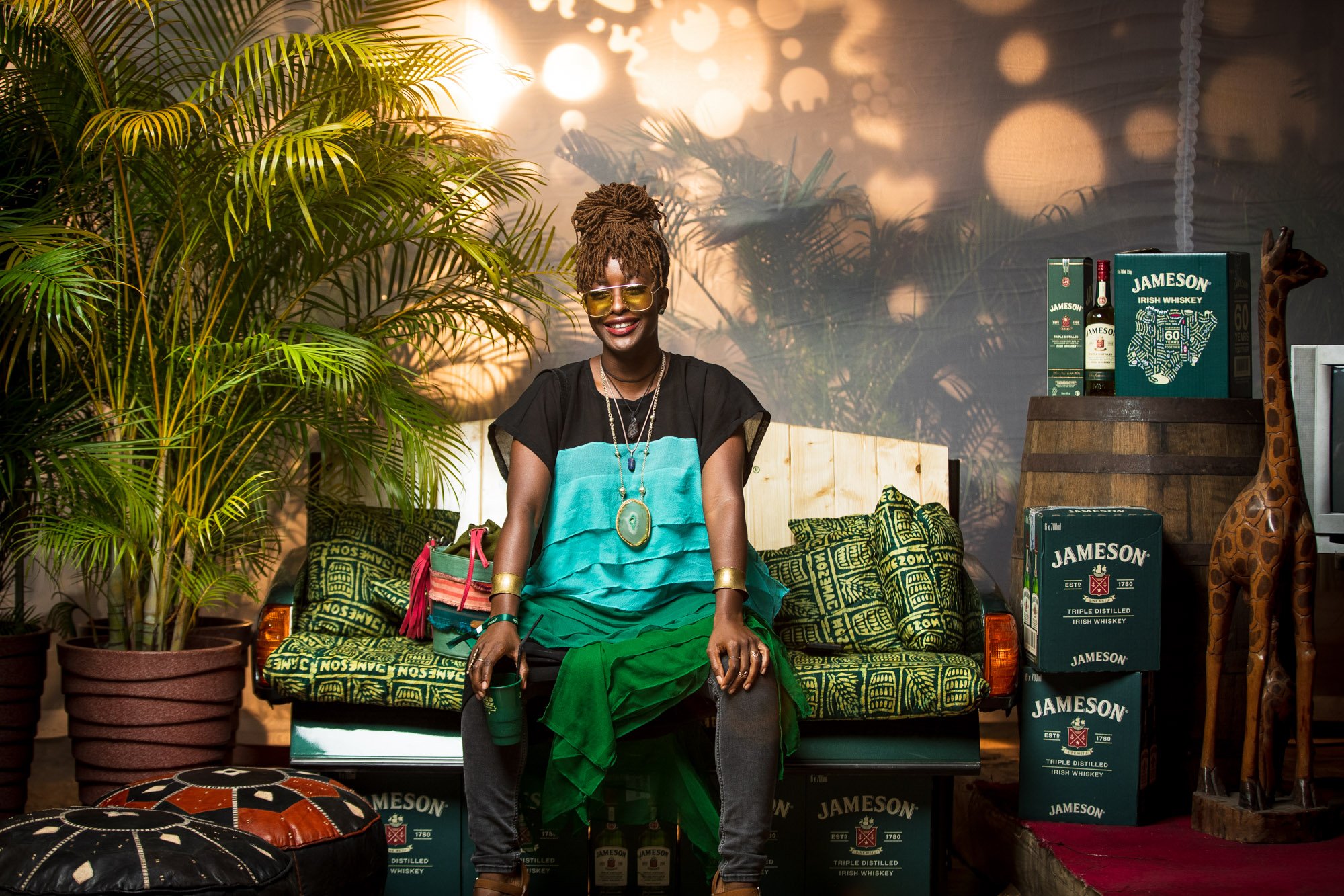 A woman with dreadlocks, wearing yellow-tinted glasses, a black top, and a turquoise and green skirt, sitting on a colorful patterned sofa in a room decorated with plants, large Jameson whiskey boxes, and a giraffe figurine, with a nature-themed mura