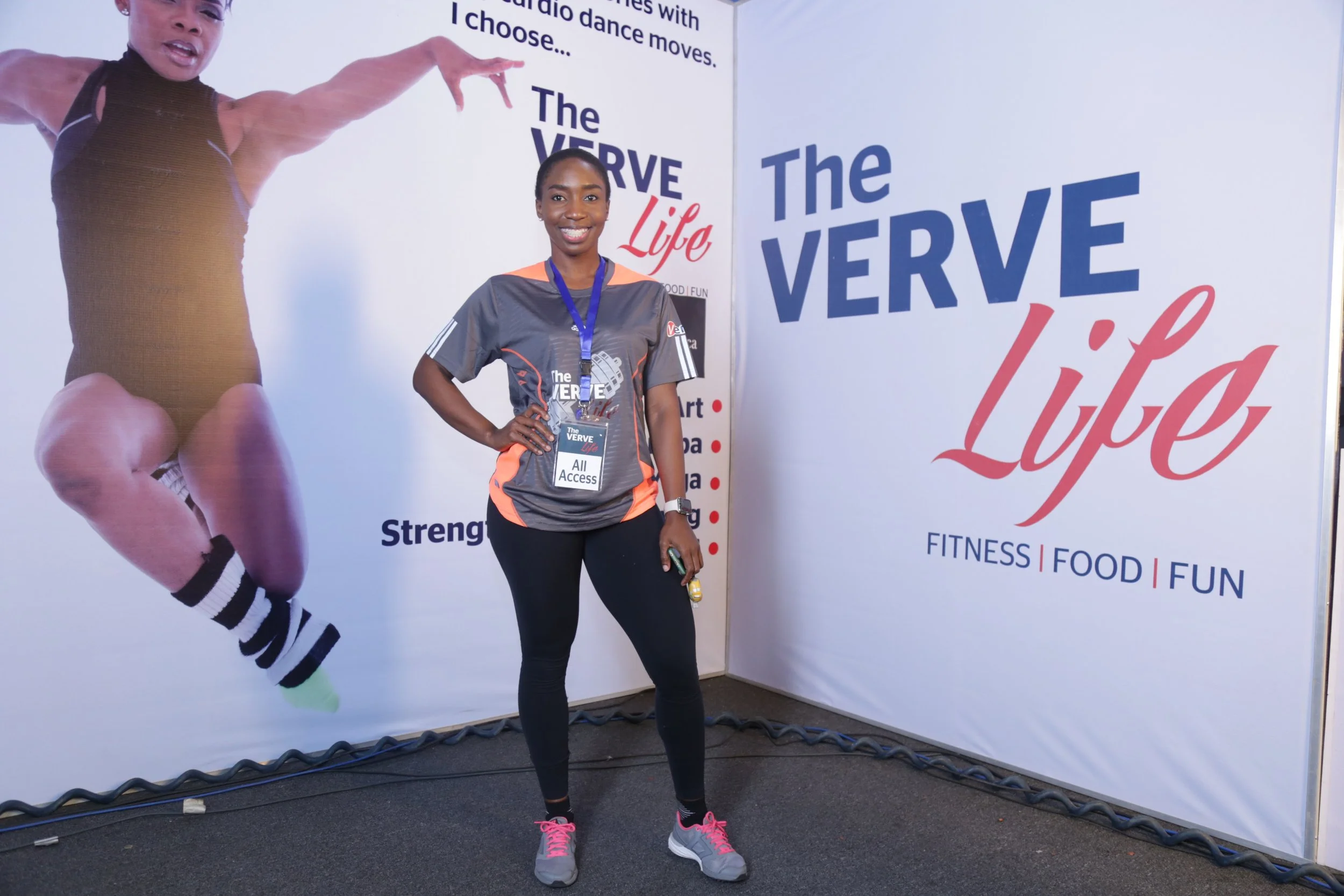 A smiling woman standing in front of a large promotional backdrop for 'The VERVE Life' fitness event. She is wearing a gray and orange athletic shirt with a medal around her neck, black leggings, and gray sneakers with pink laces. The backdrop featur