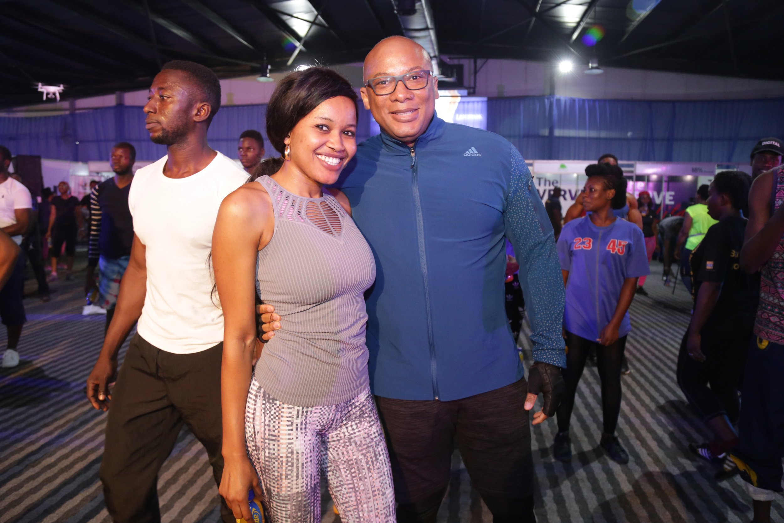 Two smiling people, a woman and a man, posing together at an indoor event with a crowd in the background.