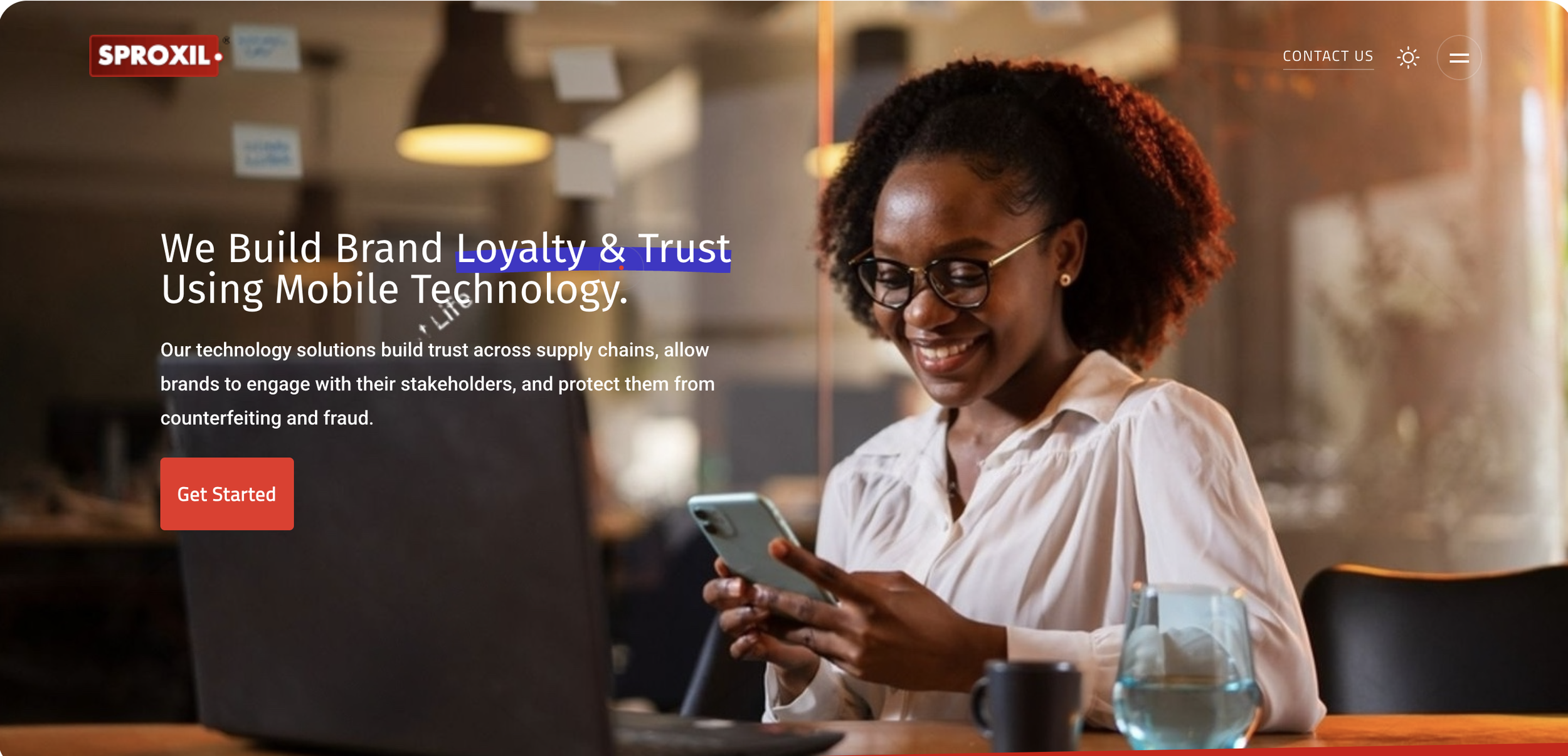 A woman with glasses and curly hair looking at her smartphone while sitting at a wooden table with a black laptop, laptop, mug, and water glass. Background shows warm lighting and a modern office cafe setting.