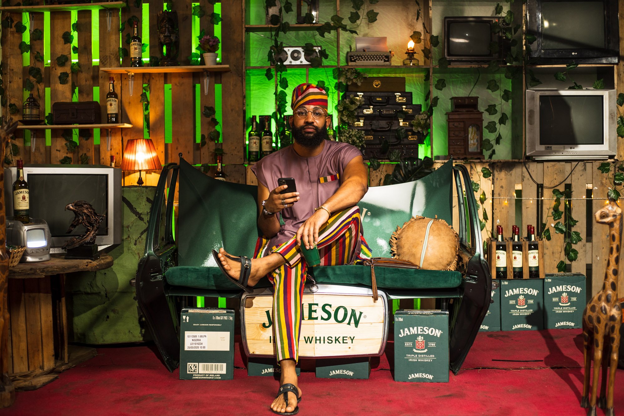 A man with a beard and glasses, dressed in a traditional African hat and colorful striped pants, sitting on a green bench in a room decorated with Jameson whiskey bottles and boxes, vintage televisions, and lush green plants, holding a smartphone.