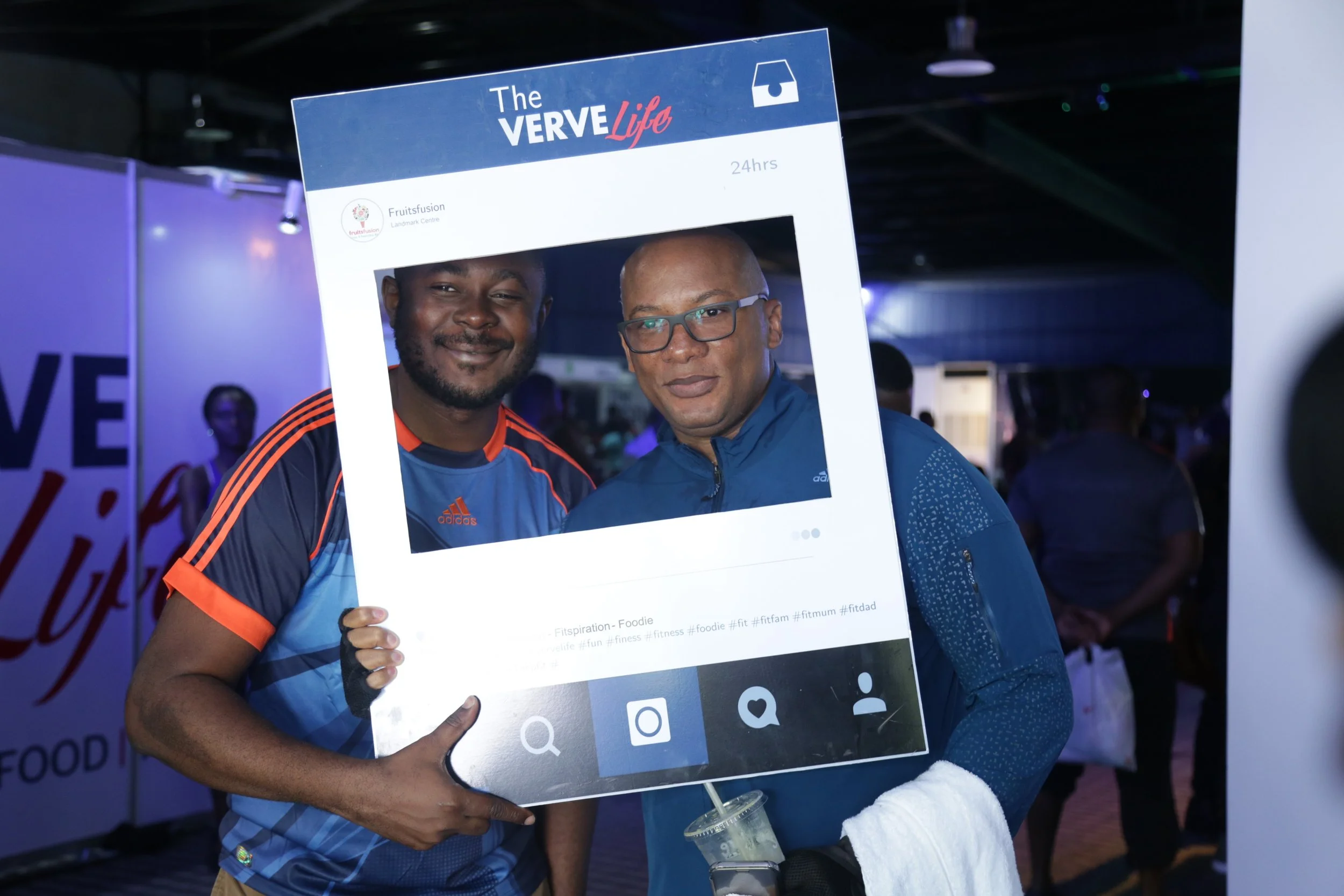 Two men at a fitness event holding a large Instagram photo frame prop. They are smiling, dressed in athletic wear, and standing in a dimly lit setting with other people in the background.