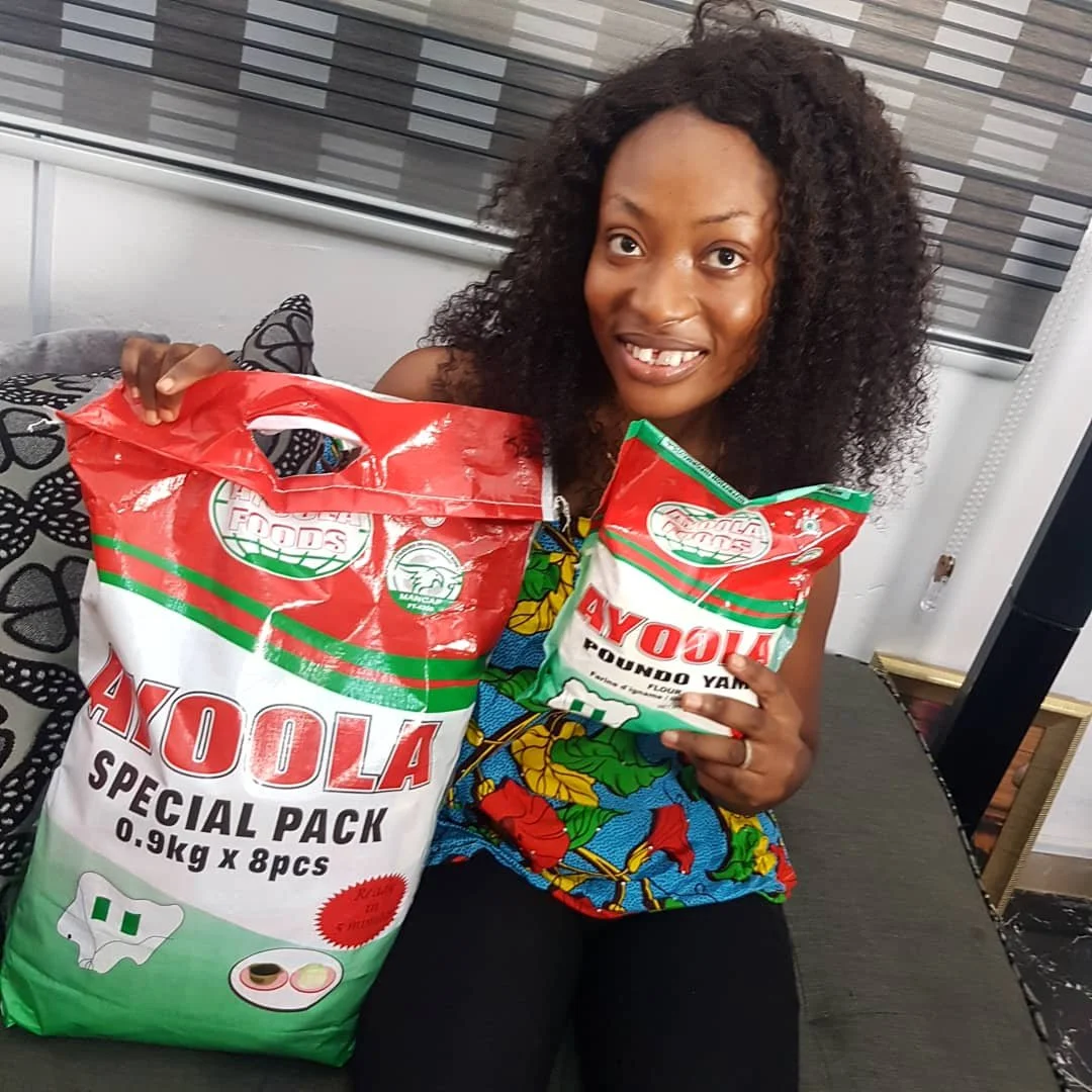 A woman with curly black hair is sitting on a sofa, holding two bags of AYOLA flour, one large and one small.