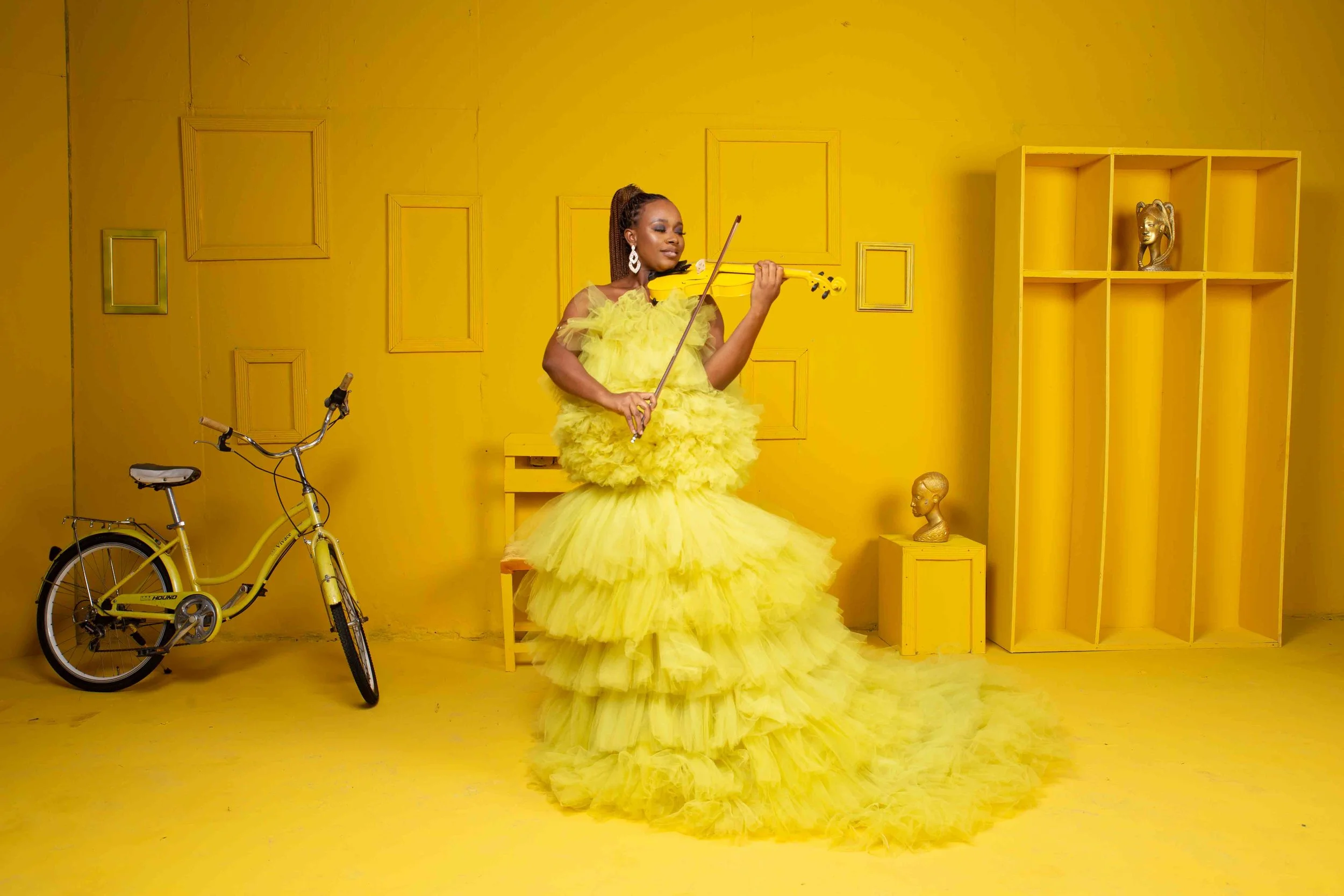 A woman in a bright yellow, ruffled, floor-length gown standing on a matching yellow floor in a yellow room, playing a yellow violin.