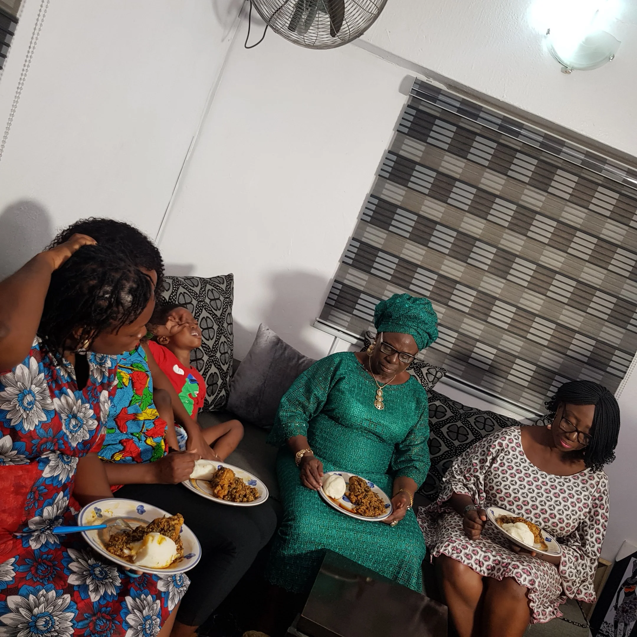 Four women and one young girl sitting on a sofa, enjoying a meal of rice, beans, and plantains, in a living room with window blinds, a ceiling fan, and a wall light.