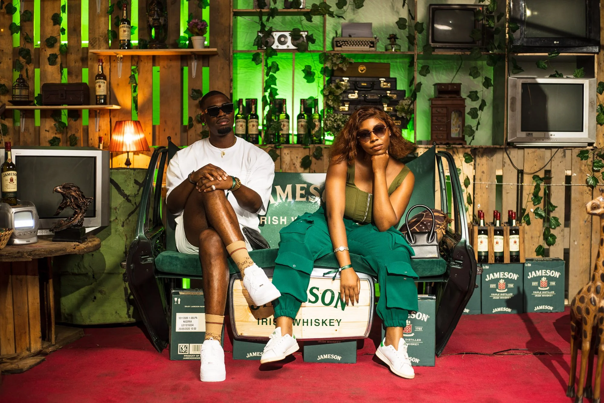 Two young adults sitting on a vintage truck decorated with Jameson Irish Whiskey branding, surrounded by shelves with bottles, radios, and TV sets, in a rustic setting with green lighting and decorative plants.