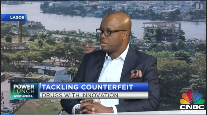 A man wearing glasses and a dark suit with a light blue shirt and handkerchief in pocket, sitting in a studio with an outdoor cityscape background of Lagos, Nigeria, discussing counterfeit drugs.