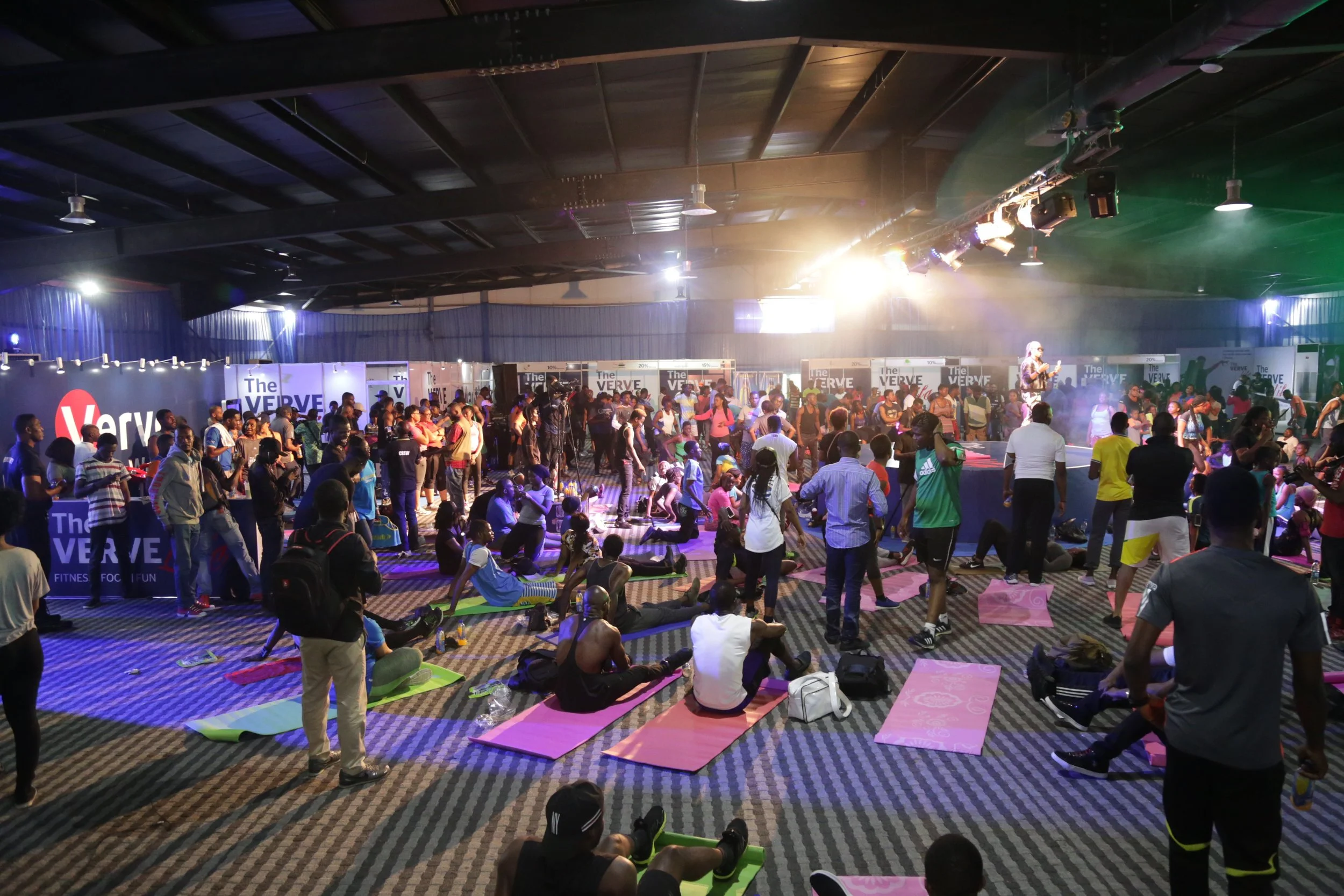 A large indoor event with many people participating in a fitness class or dance session, some on yoga mats, others standing or sitting, with a stage in the background featuring a performer and banners reading "The VERVE."