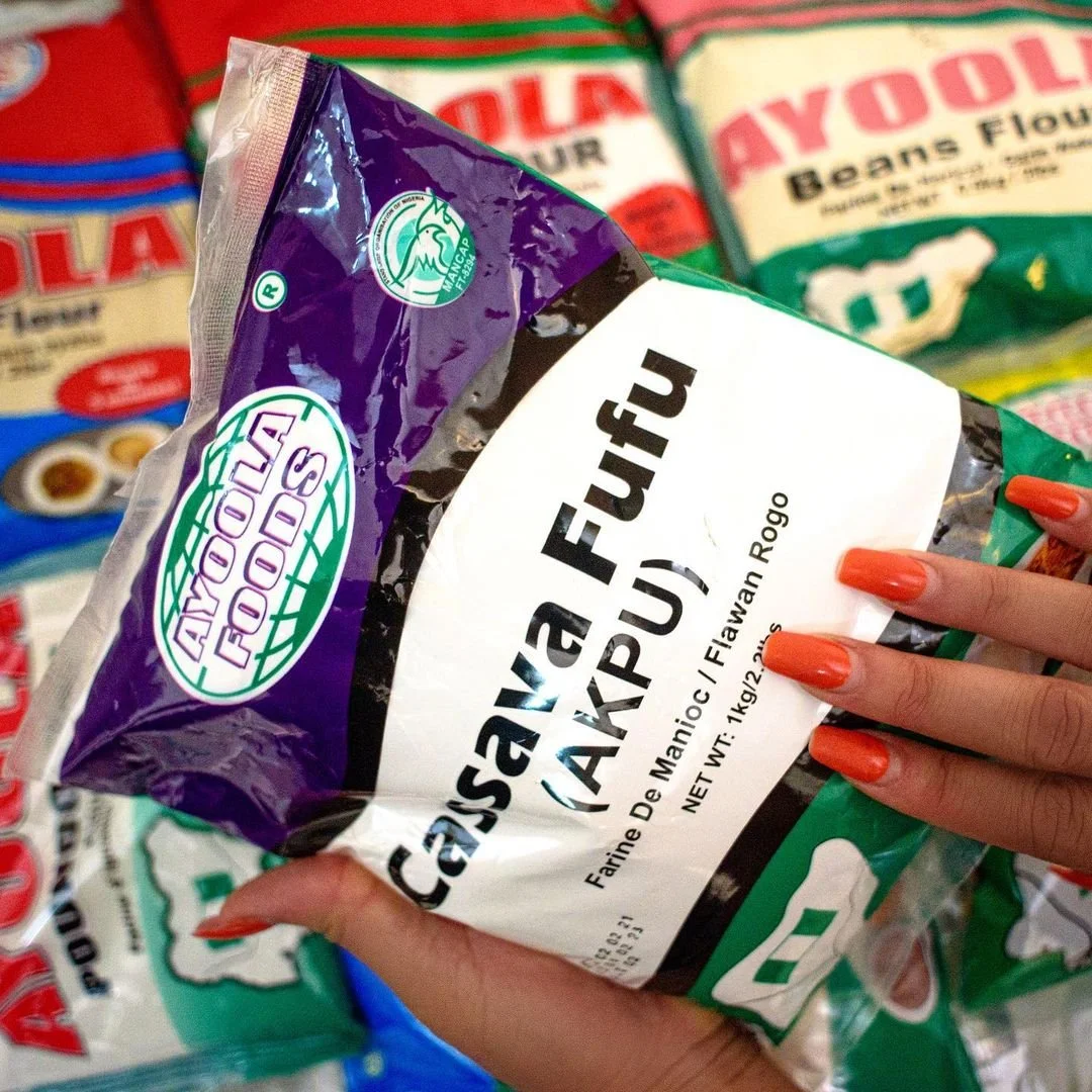 Close-up of a person holding a bag of cassava flour from Ayoola Foods, with other flour bags visible in the background.
