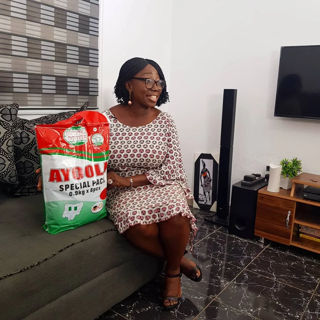 A woman sitting on a sofa in a living room, smiling, with a large pack of Ayool dog food next to her. She has short curly hair, glasses, and is wearing a patterned dress and black sandals. There is a TV and entertainment unit with a plant and decorat