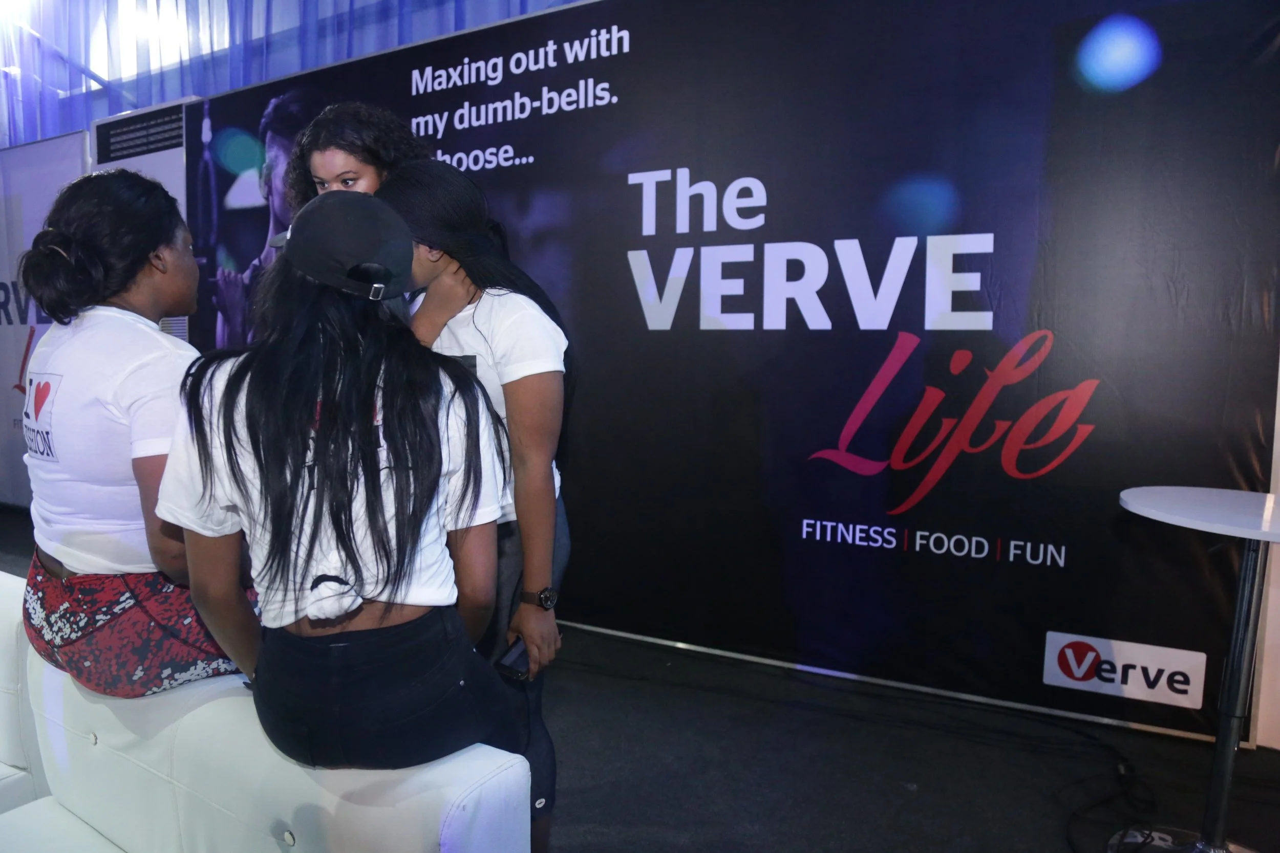 Three women sitting and talking at an event booth with a large black banner that reads "The VERVE Life" and mentions fitness, food, and fun.