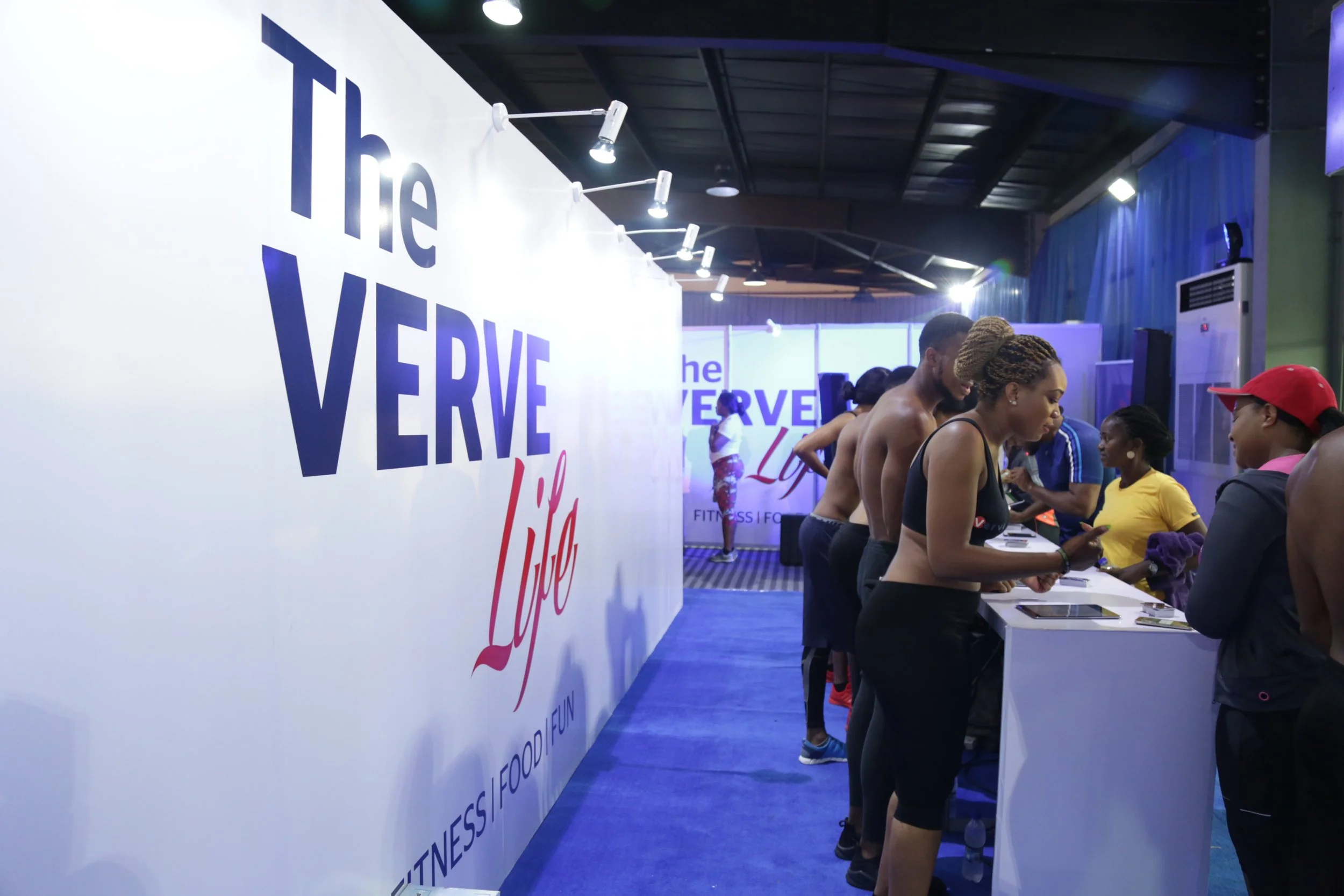 People gather at a booth with a large sign that reads "The VERVE Life" and a tagline "FITNESS, FOOD, FUN."