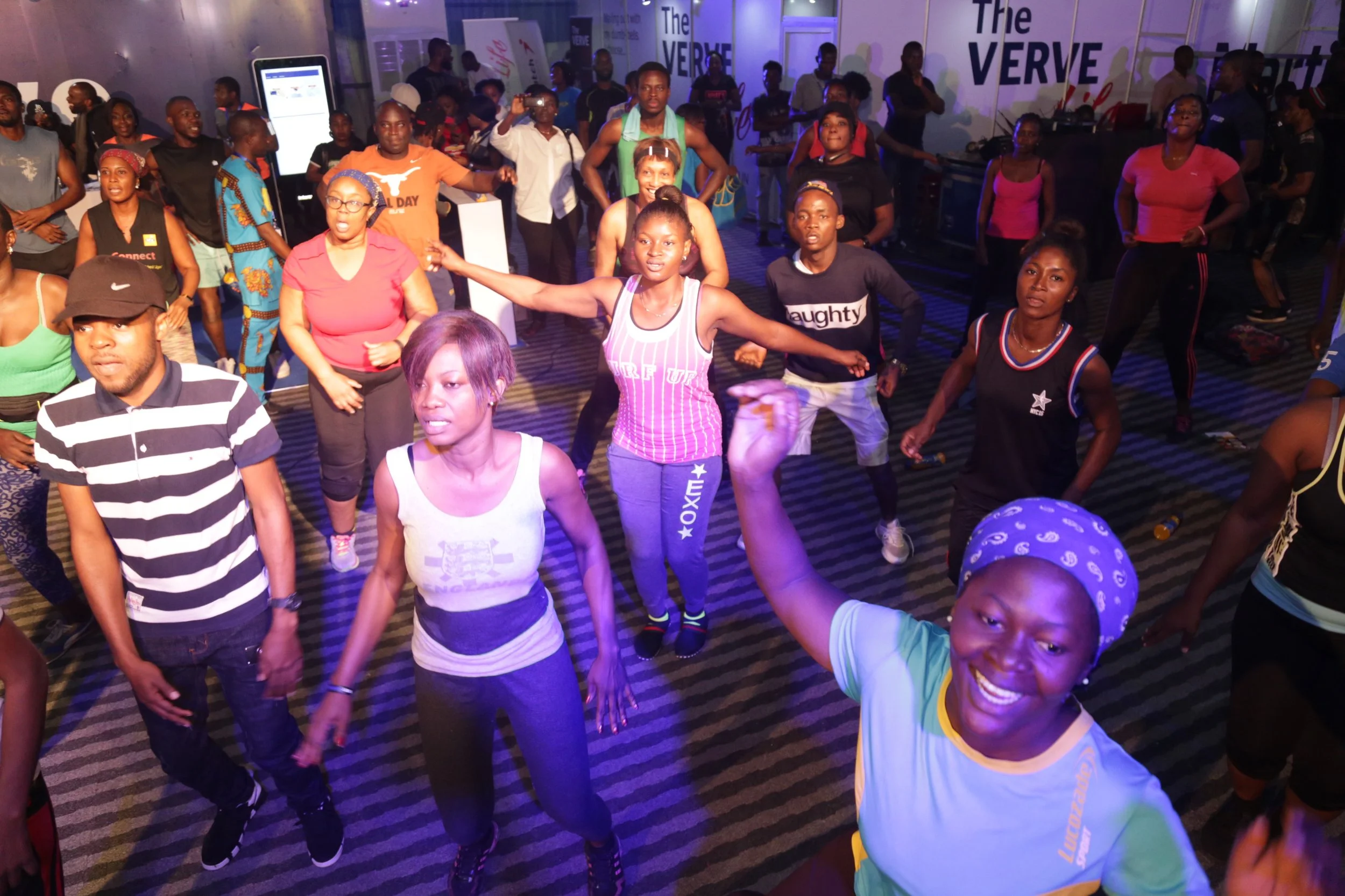 A group of people participating in a dance or fitness class indoors, with a woman at the front smiling and wearing a blue bandana. The background features a purple-lit room with banners and other participants dancing.