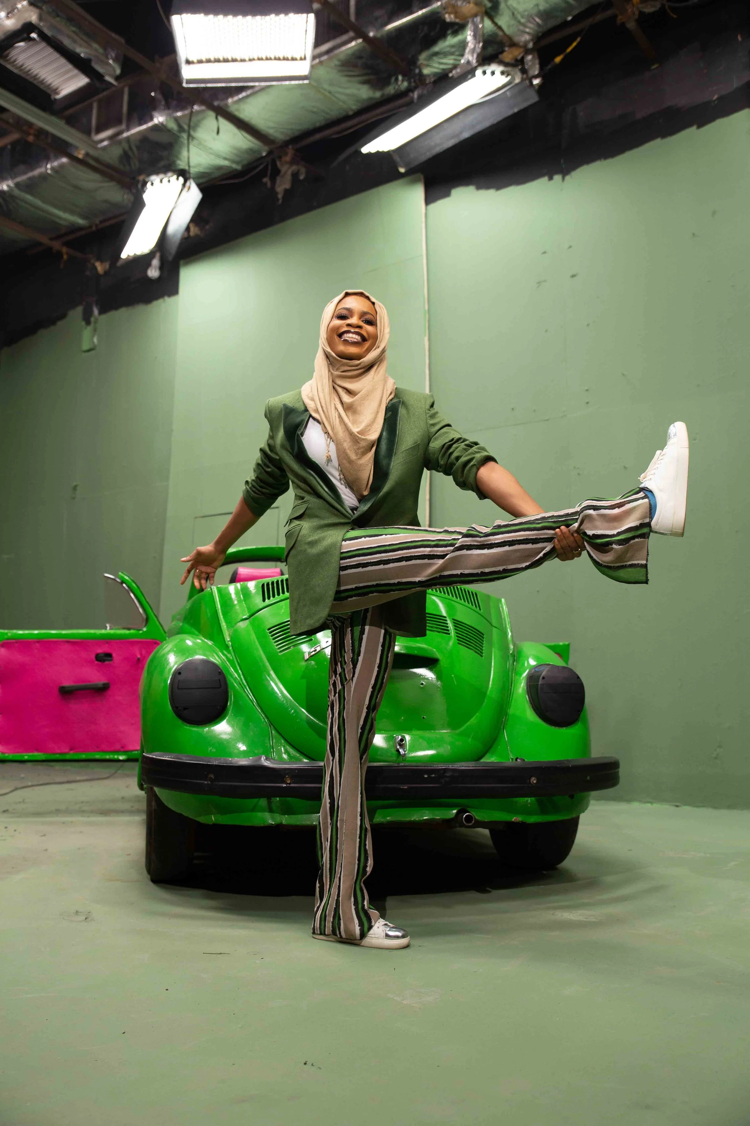 A woman dressed in a blazer and striped pants, smiling and stretching her leg, standing in front of a green vintage car inside a room with green walls and fluorescent lights.