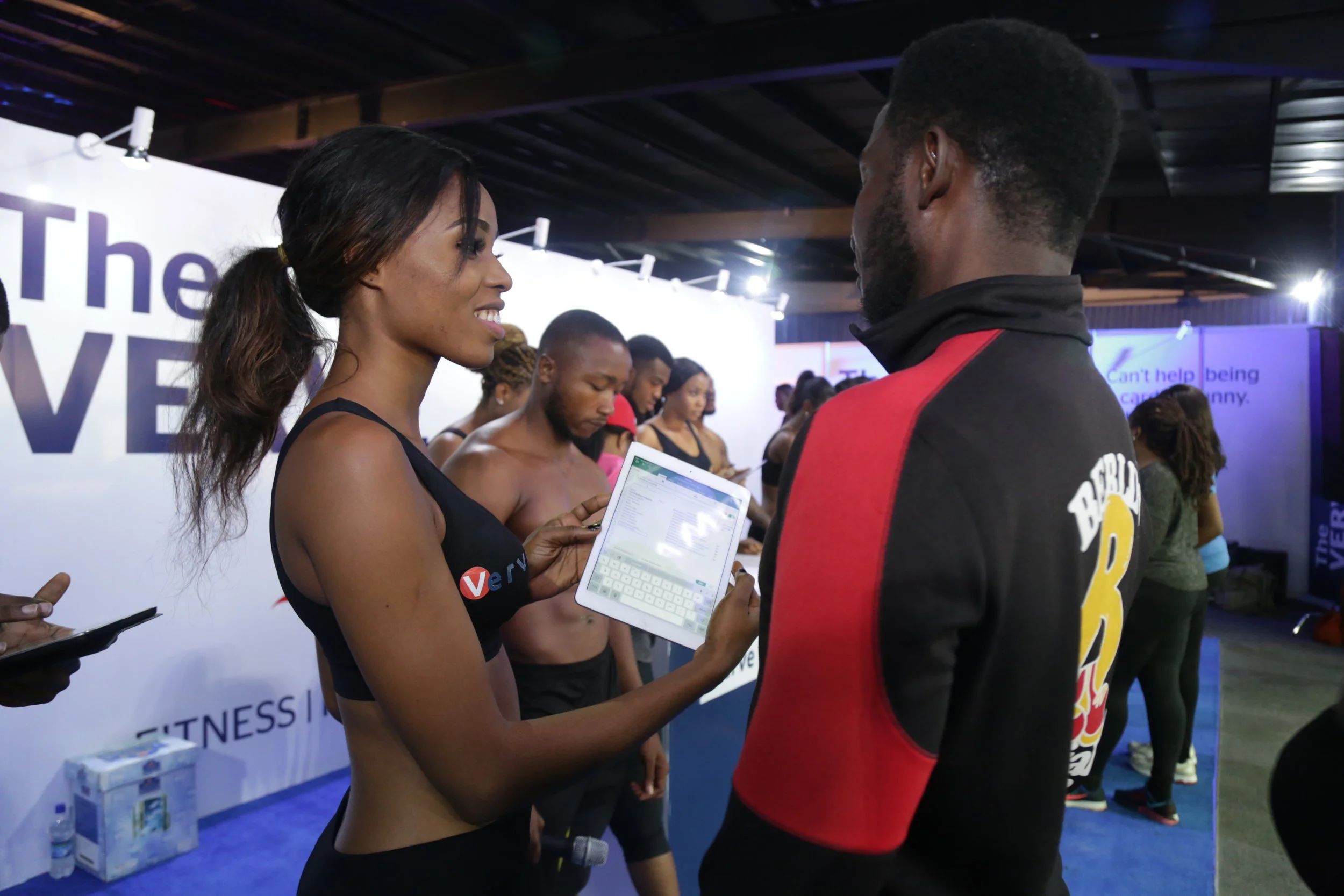 A woman in fitness attire holding a tablet, talking to a man in a black and red jacket, with a line of people in athletic clothing in the background at a fitness event.