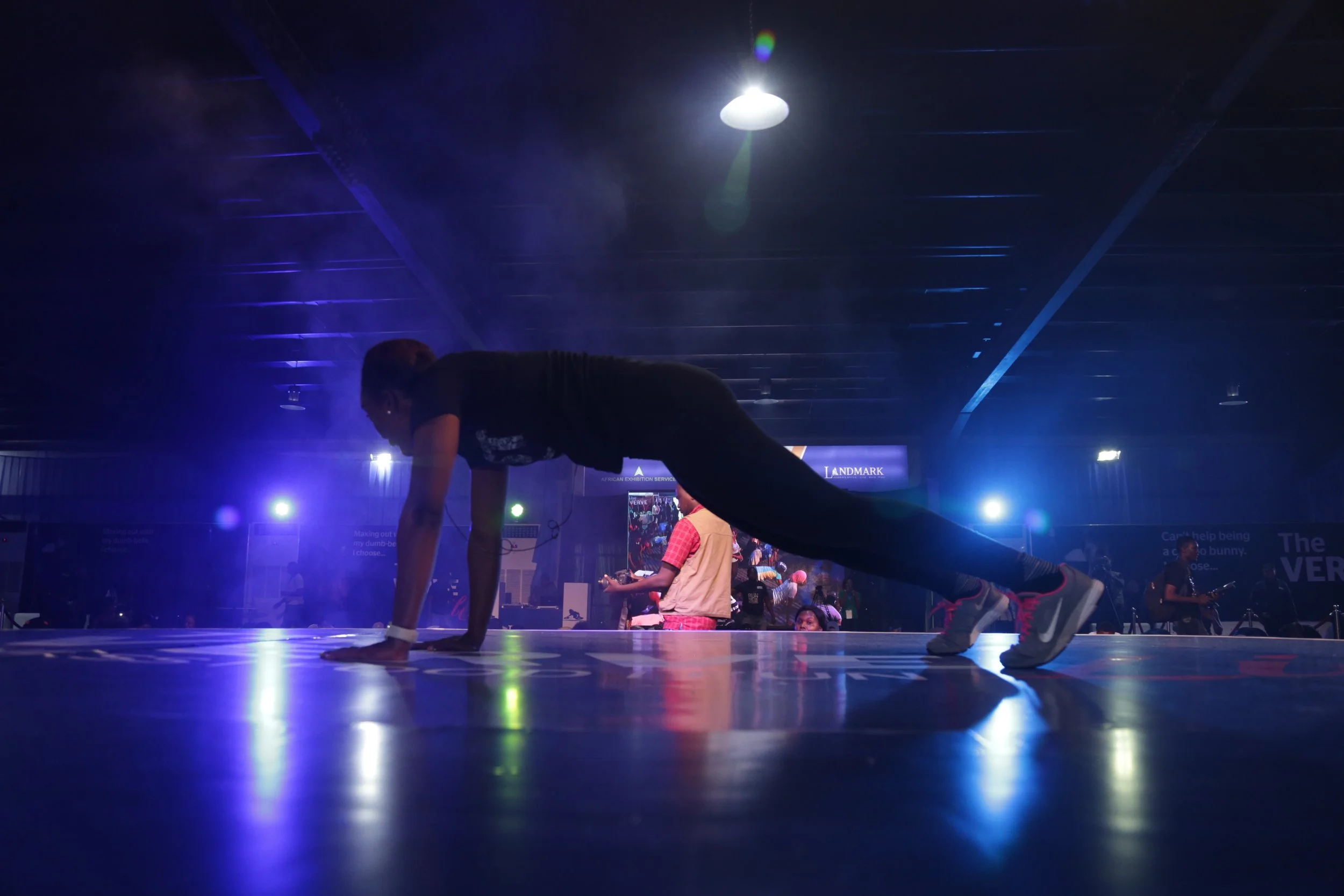 A person is in a push-up position on a stage or floor with dark lighting and spotlights in the background.