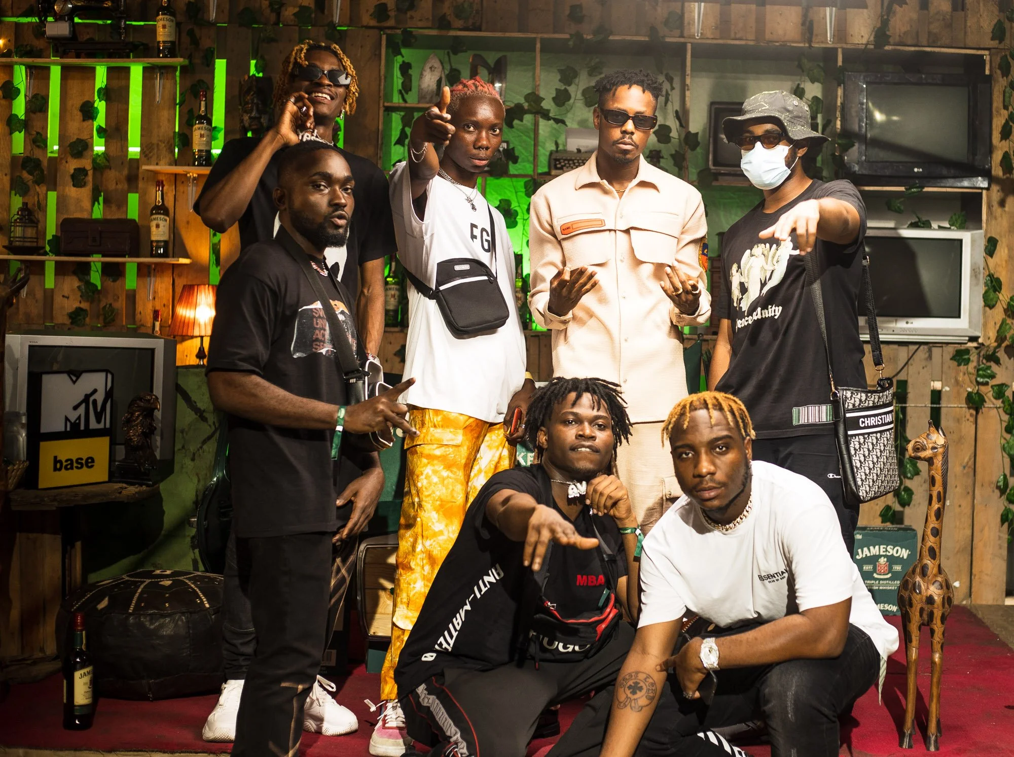 A group of ten young men posing together in a room decorated with wooden walls, green lighting, plants, and various decor items including a giraffe statue, TV, and posters. Some are standing, some kneeling or sitting, dressed casually, with one weari