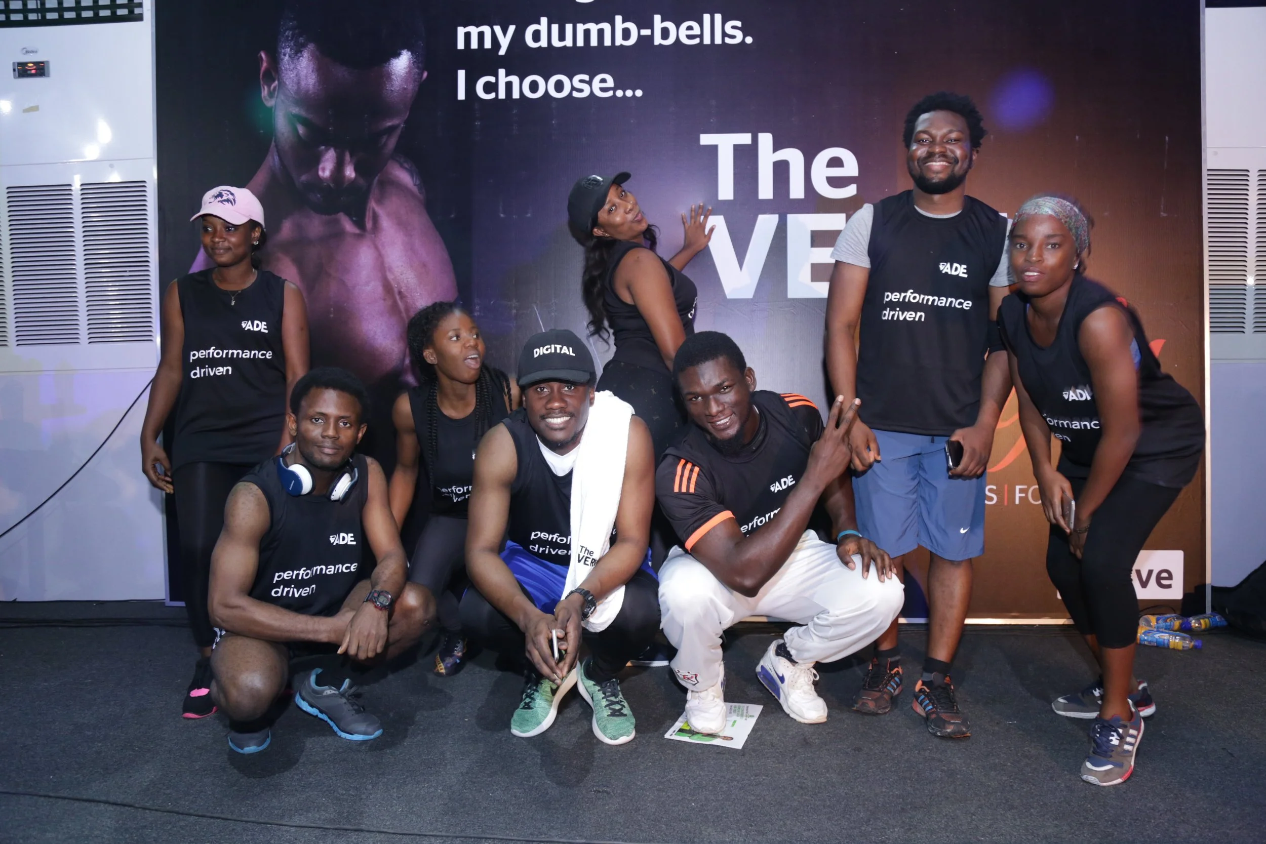 Group of nine people posing for a photo in front of a promotional backdrop with the message "my dumb-bells. I choose... The VE". The group is wearing black shirts with the words "performance driven" and some are smiling and making gestures.