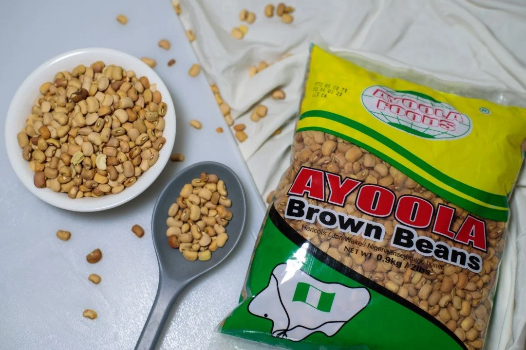 A bag of Ayoola brown beans, a bowl of beans, and a spoon with some beans on it on a light-colored surface.