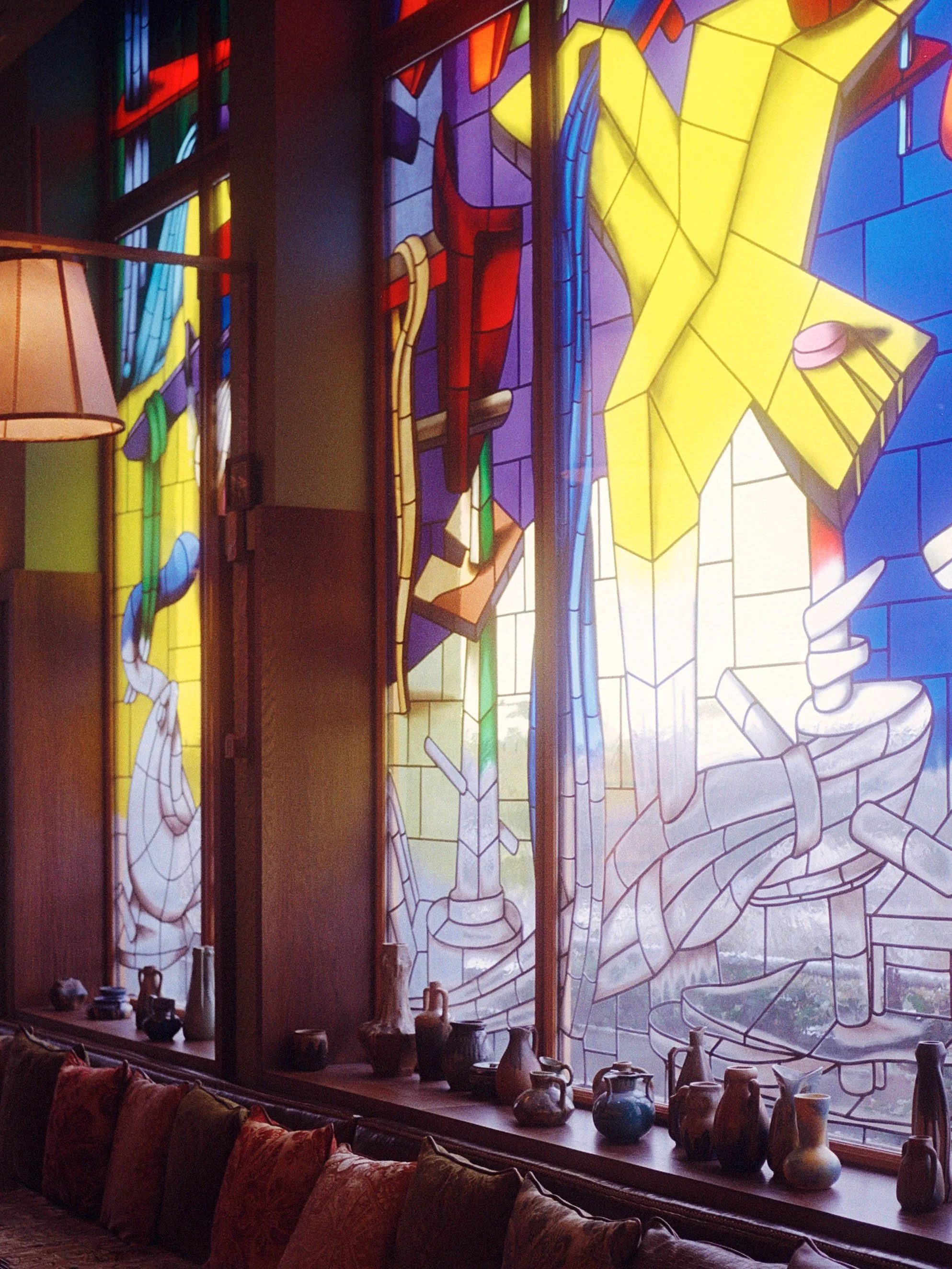 Vitrail coloré représentant une scène en style artistique abstrait, avec des formes géométriques et des couleurs vives, dans un intérieur avec des objets en poterie sur le rebord de la fenêtre.