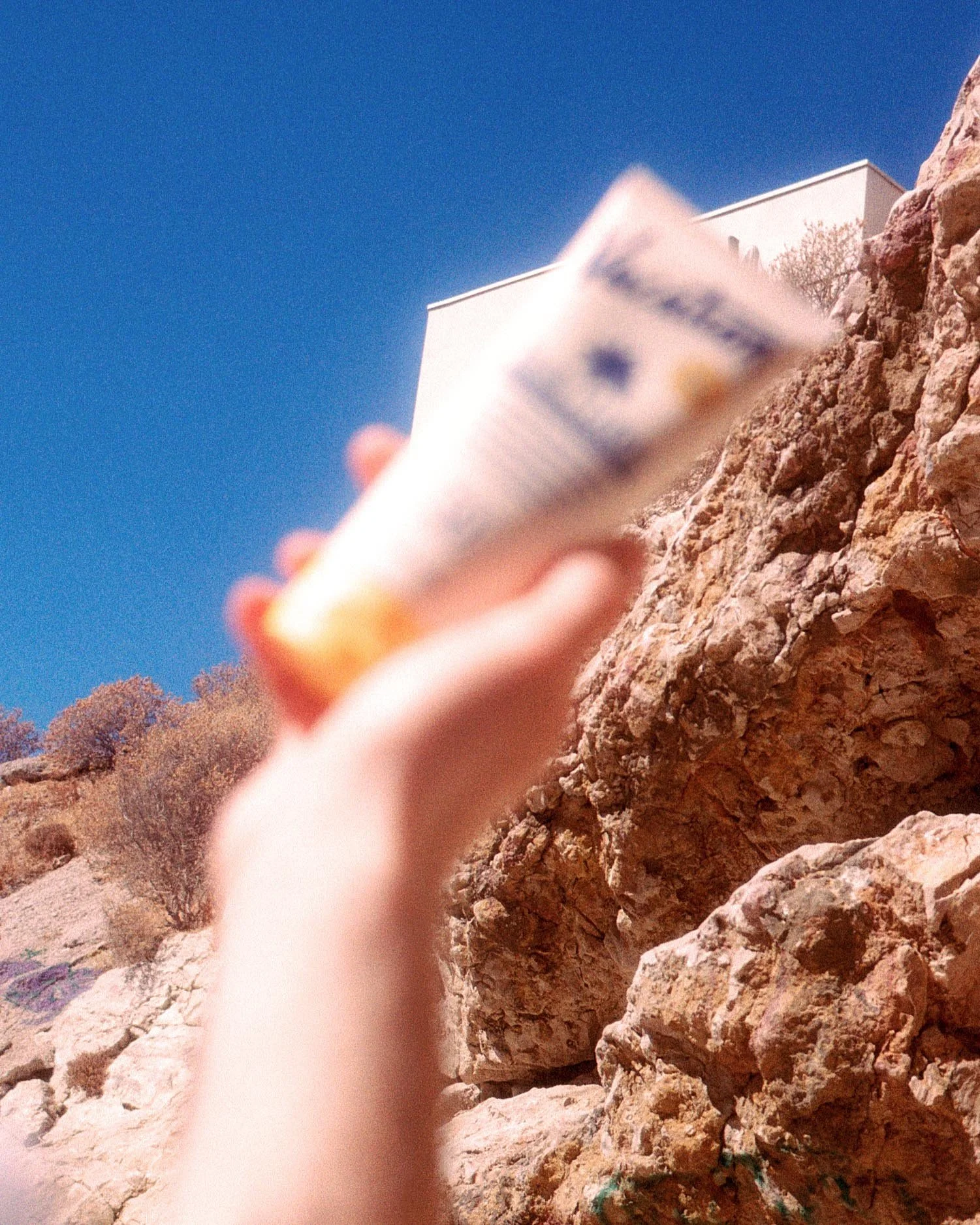 Main pointant un tube de crème solaire vers le ciel bleu avec des rochers et des arbustes en arrière-plan