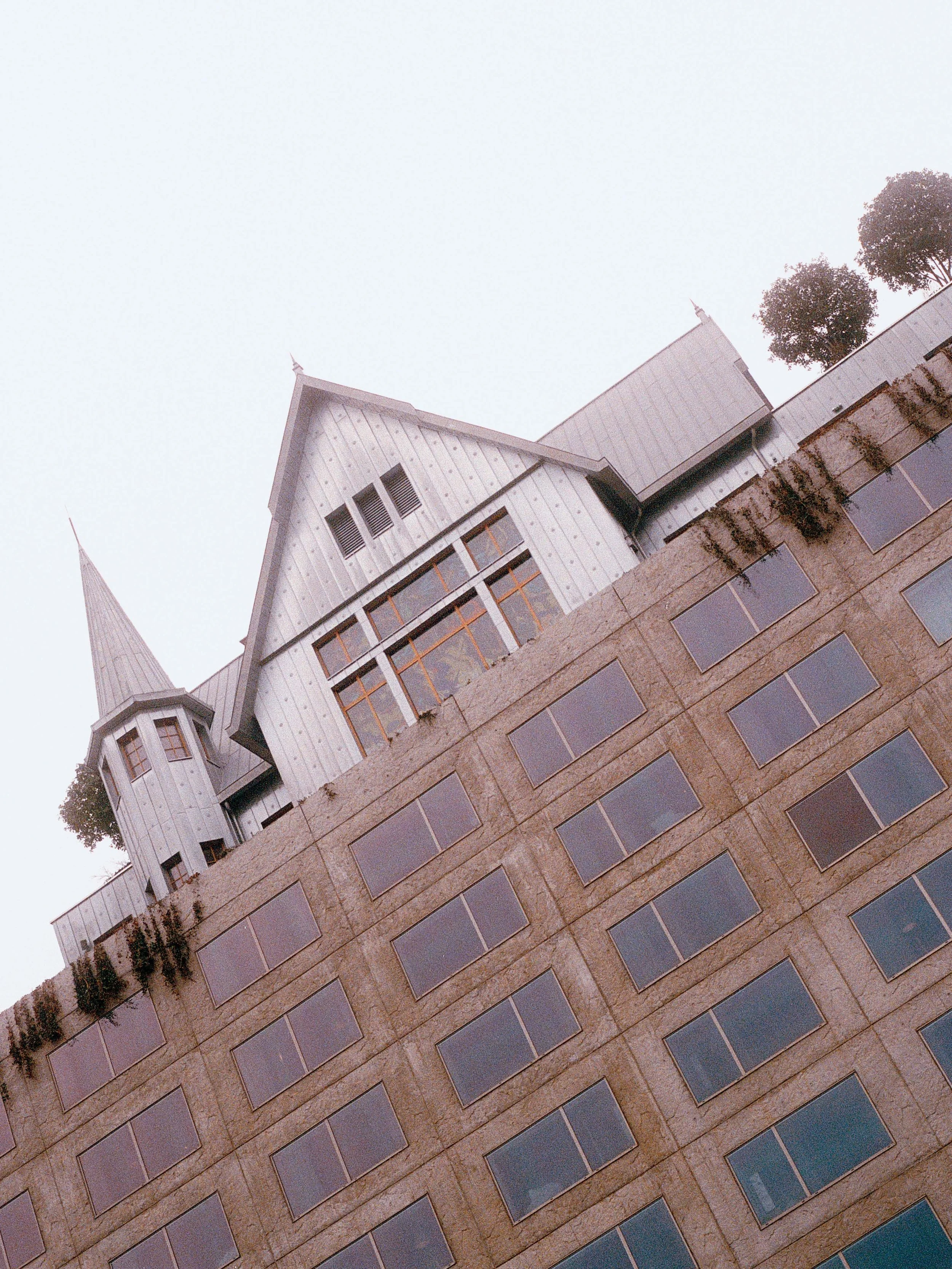 Une photo du bâtiment de la maison Heler avec plusieurs fenêtres rectangulaires, une partie en pierre et une autre en métal, avec des arbres en arrière-plan et un ciel clair.