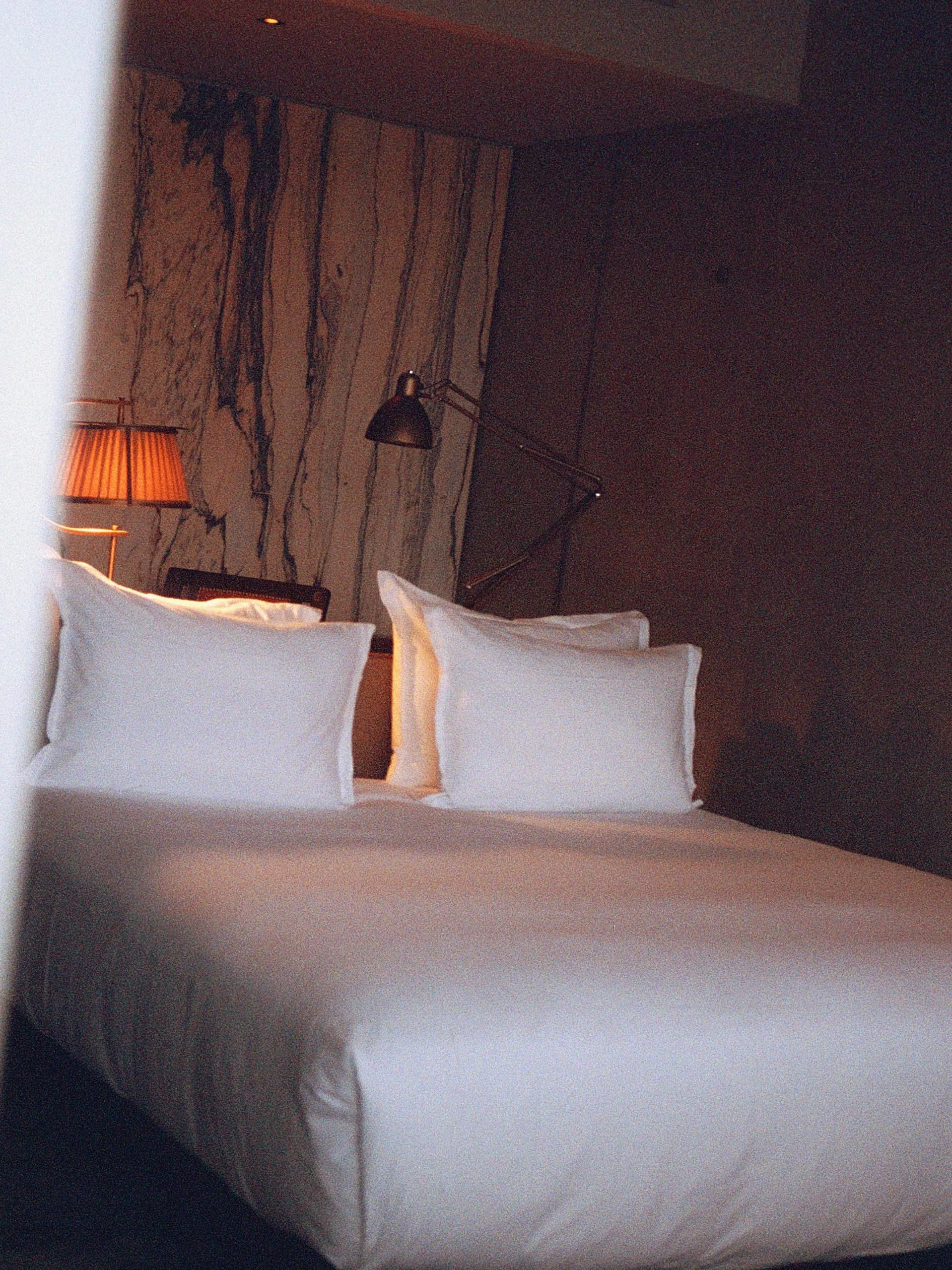 Un lit avec des draps blancs et deux oreillers, dans une chambre avec un mur en marbre, une lampe de chevet et un lampadaire