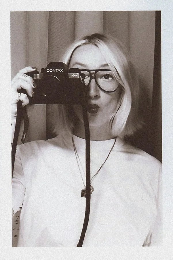 Une jeune femme blonde porte des lunettes, prend une photo d'elle-même avec un appareil photo argentique contax, et fait un visage souriant ou surpris. Elle porte un t-shirt blanc et une chaîne avec un pendentif, avec une décoration simple en arrière-plan.