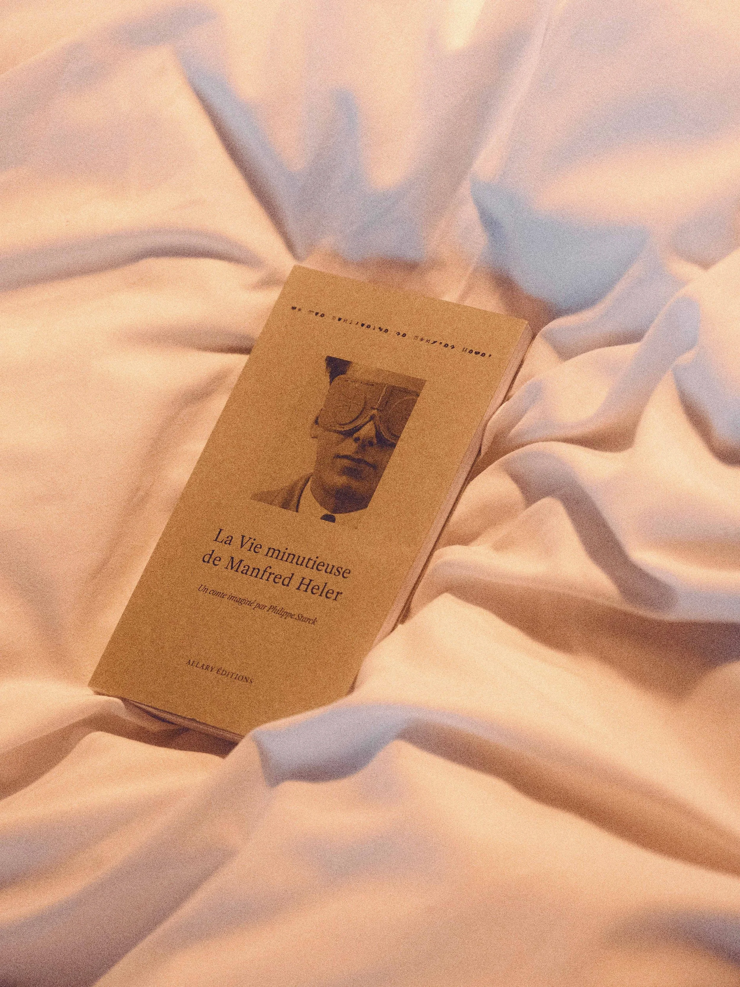 Un livre intitulé 'La Vie minutieuse de Manfred Heler' posé sur des draps blancs. La couverture montre un portrait en noir et blanc d'un homme portant des lunettes. Dans la chambre de la maison heler