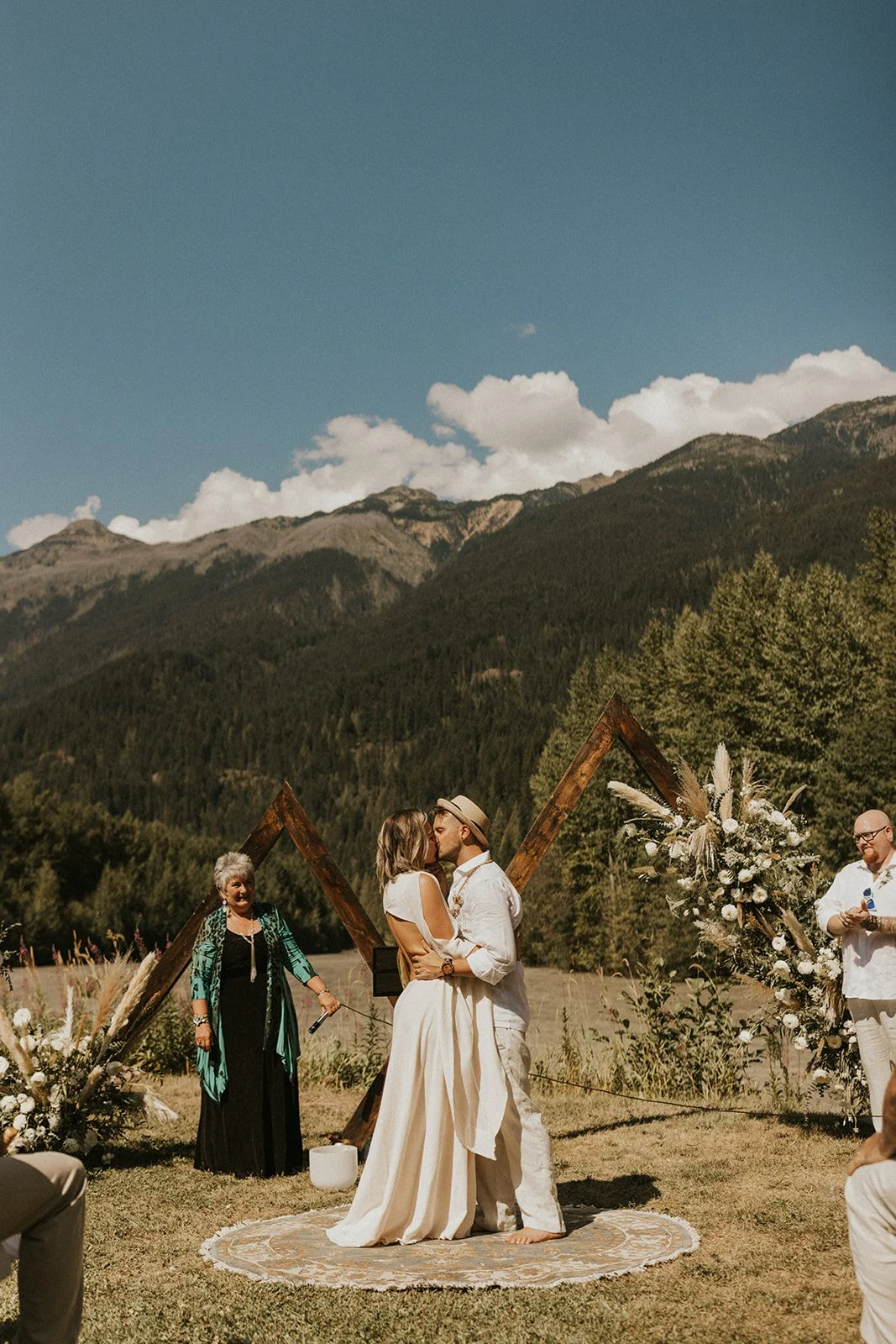 A couple kisses during an outdoor wedding ceremony with mountainous landscape in the background, a wooden arch decorated with white flowers and greenery, and a circular rug under the couple.