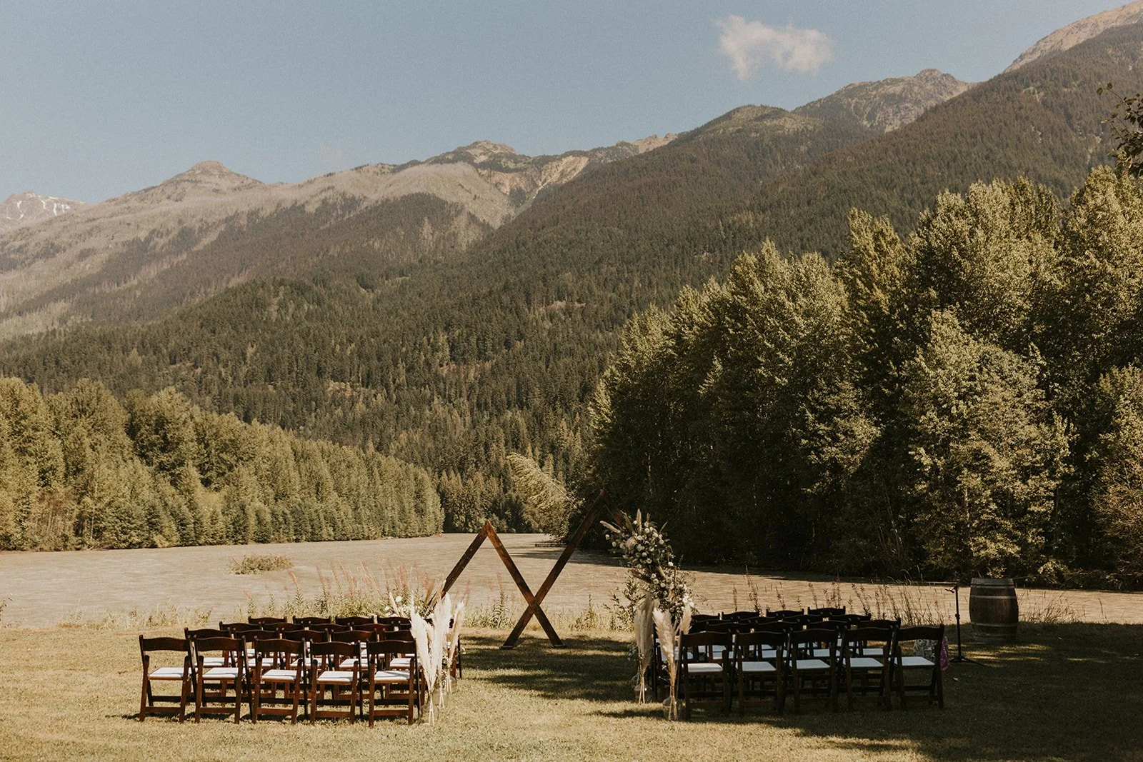 Outdoor wedding setup with rows of chairs, floral decorations, a wooden arch, and scenic mountain and forest view in the background.
