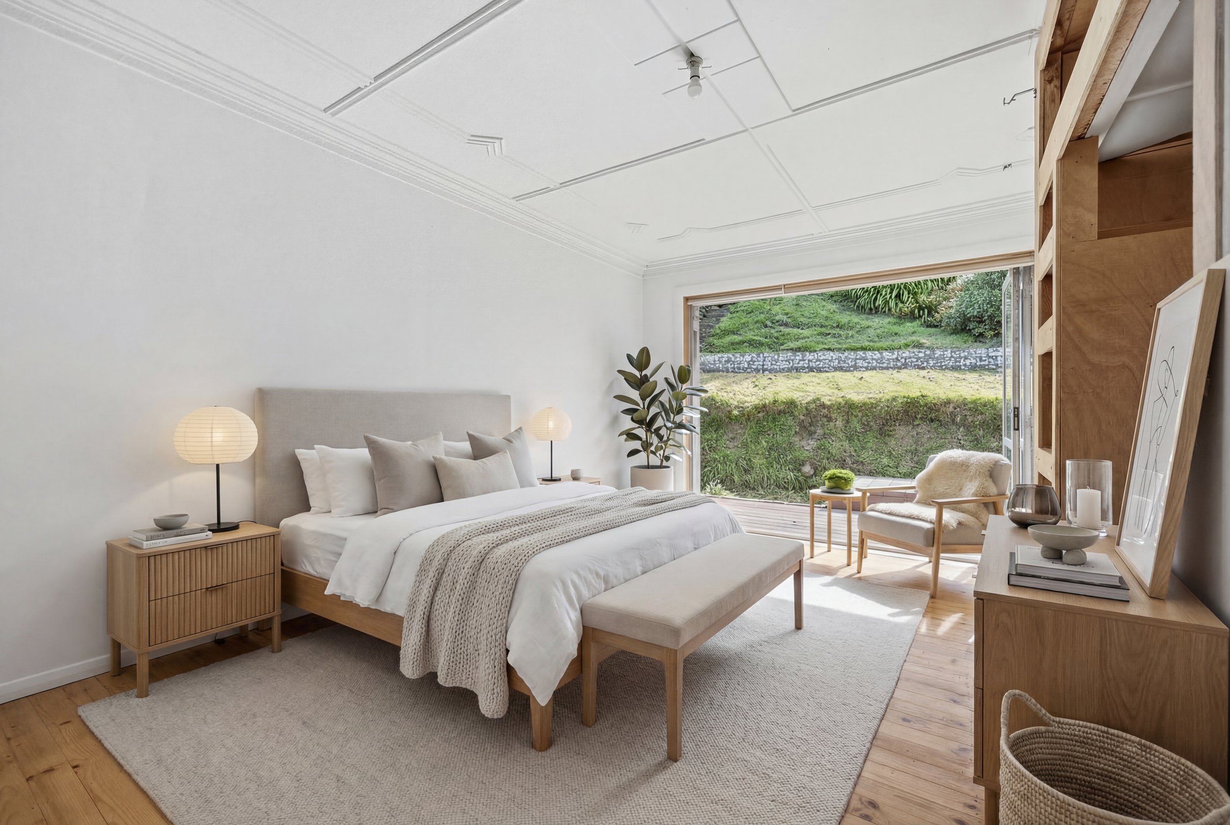 Clean and modern bedroom with beige upholstered bed, white bedding, black decorative pillow, bedside tables with lamps, large window with green curtains, potted plant, and geometric area rug.