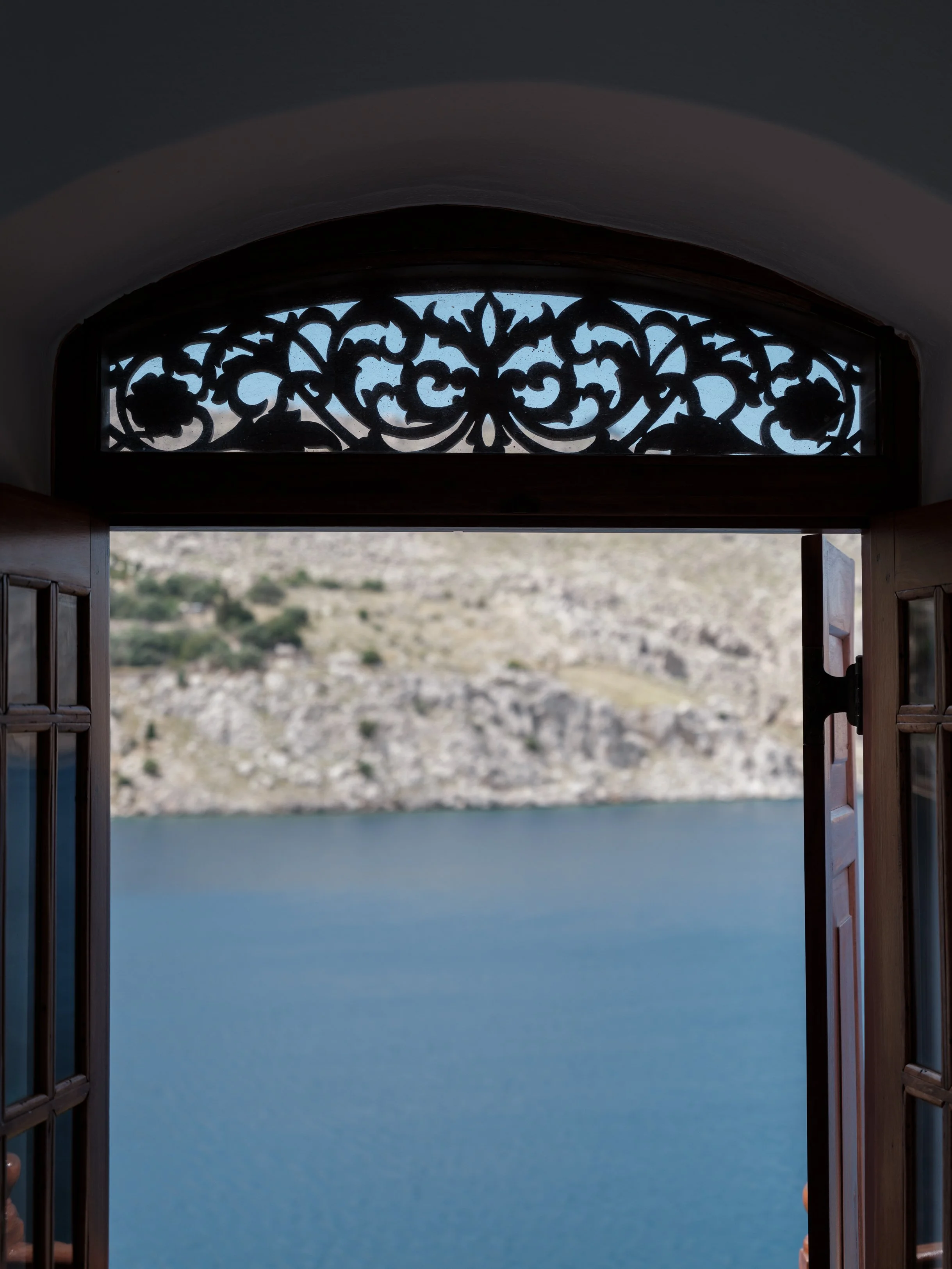 Eos suite view through a decorative wooden window frame overlooking nimborio bay and a distant landscape with rocky mountains at the selene greece, symi