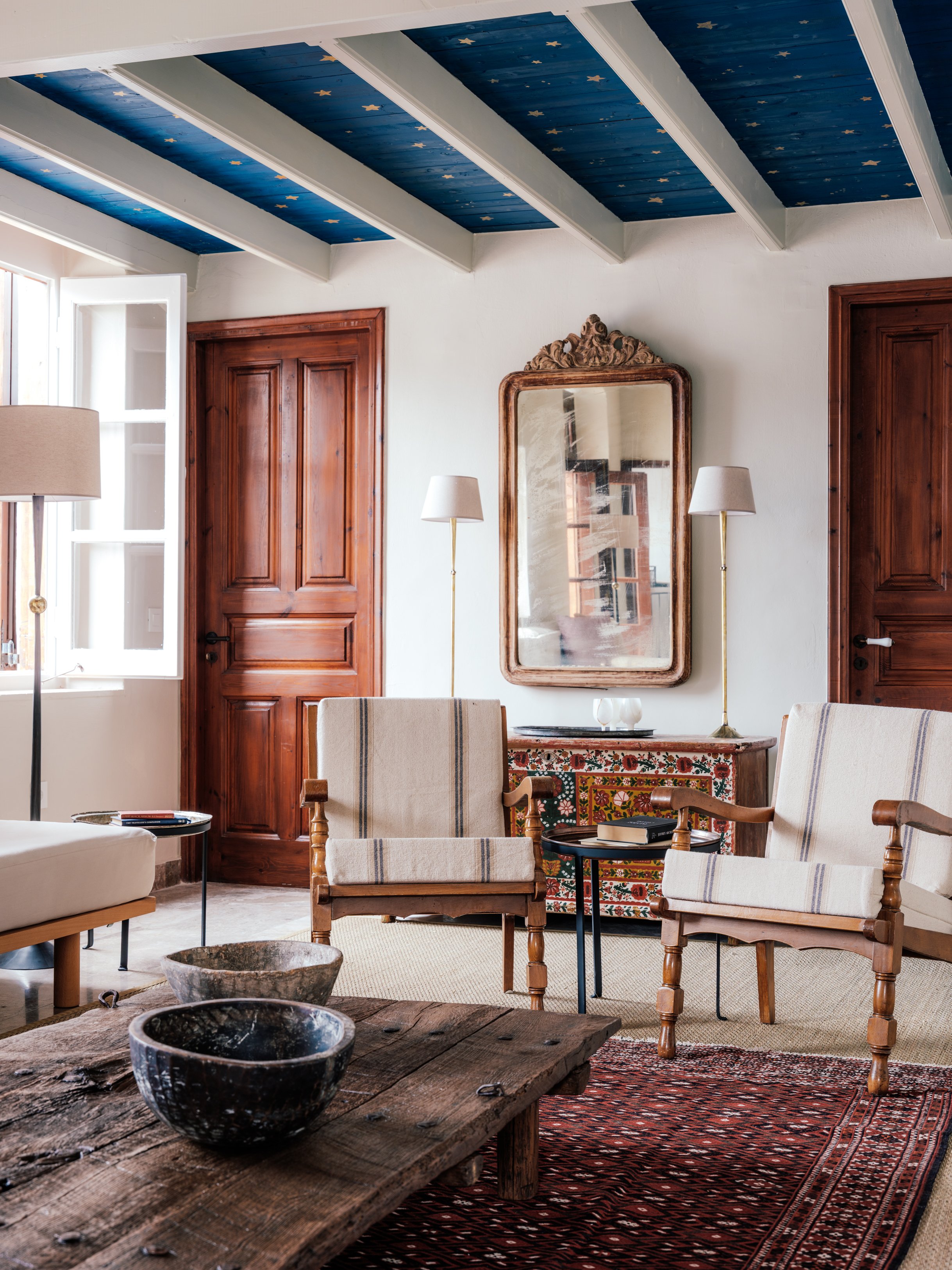 Living room with wooden furniture, a large mirror with decorative top, two floor lamps, wooden doors, a window, and a patterned rug at The Selene Greece, Symi