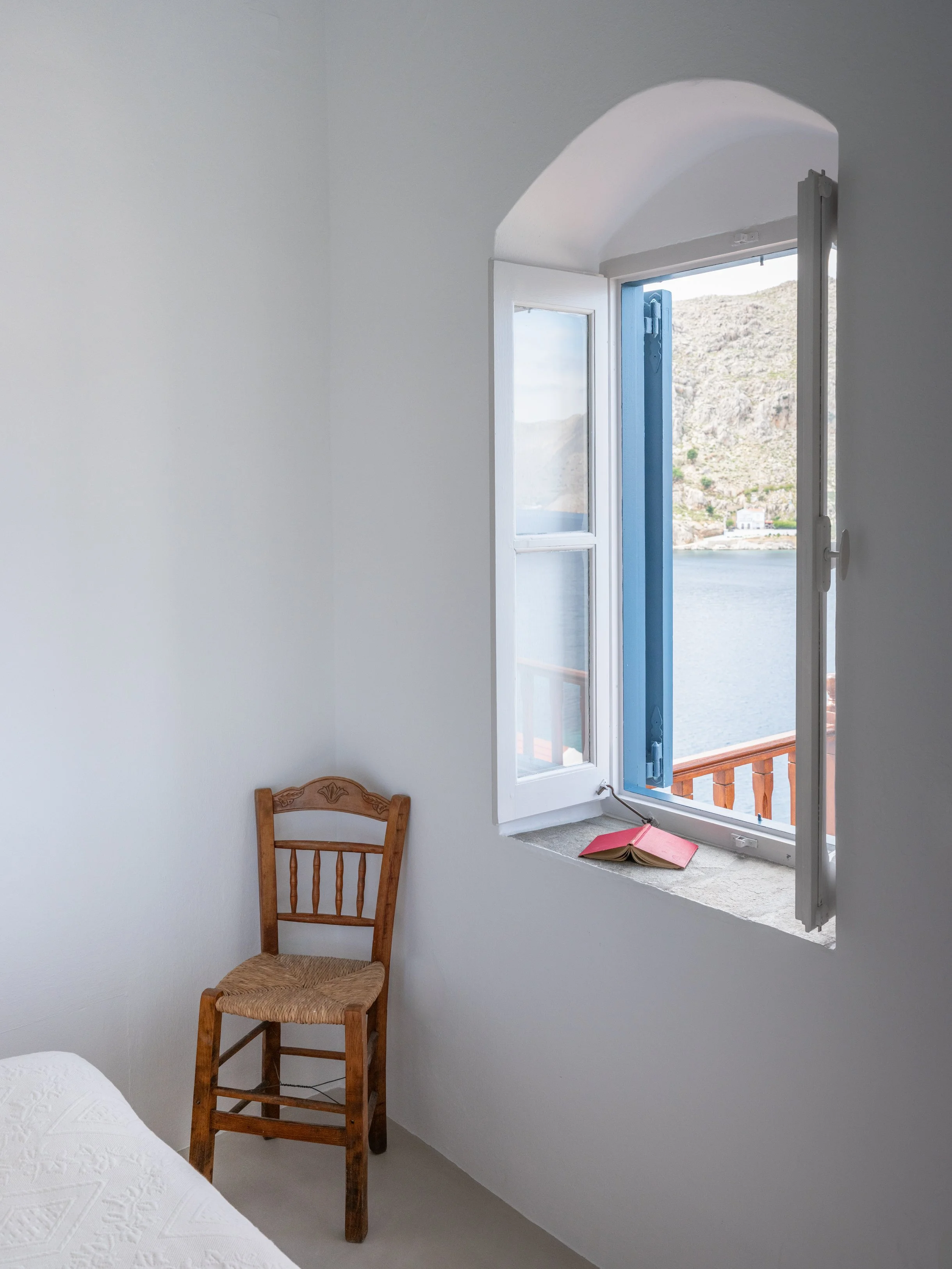 Eos bedroom with white walls and an open window overlooking the sea Nimborio bay and mountains. A wooden chair with woven seat at The Selene Greece, Symi.